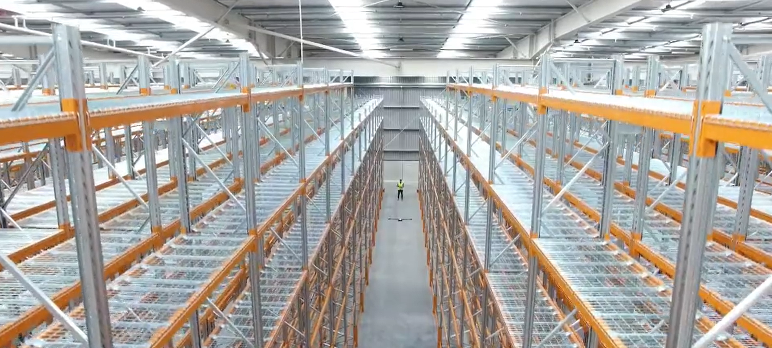 A large empty warehouse or storage facility with tall metal shelves and orange supports, with a single person in a high-visibility vest standing in the middle aisle.