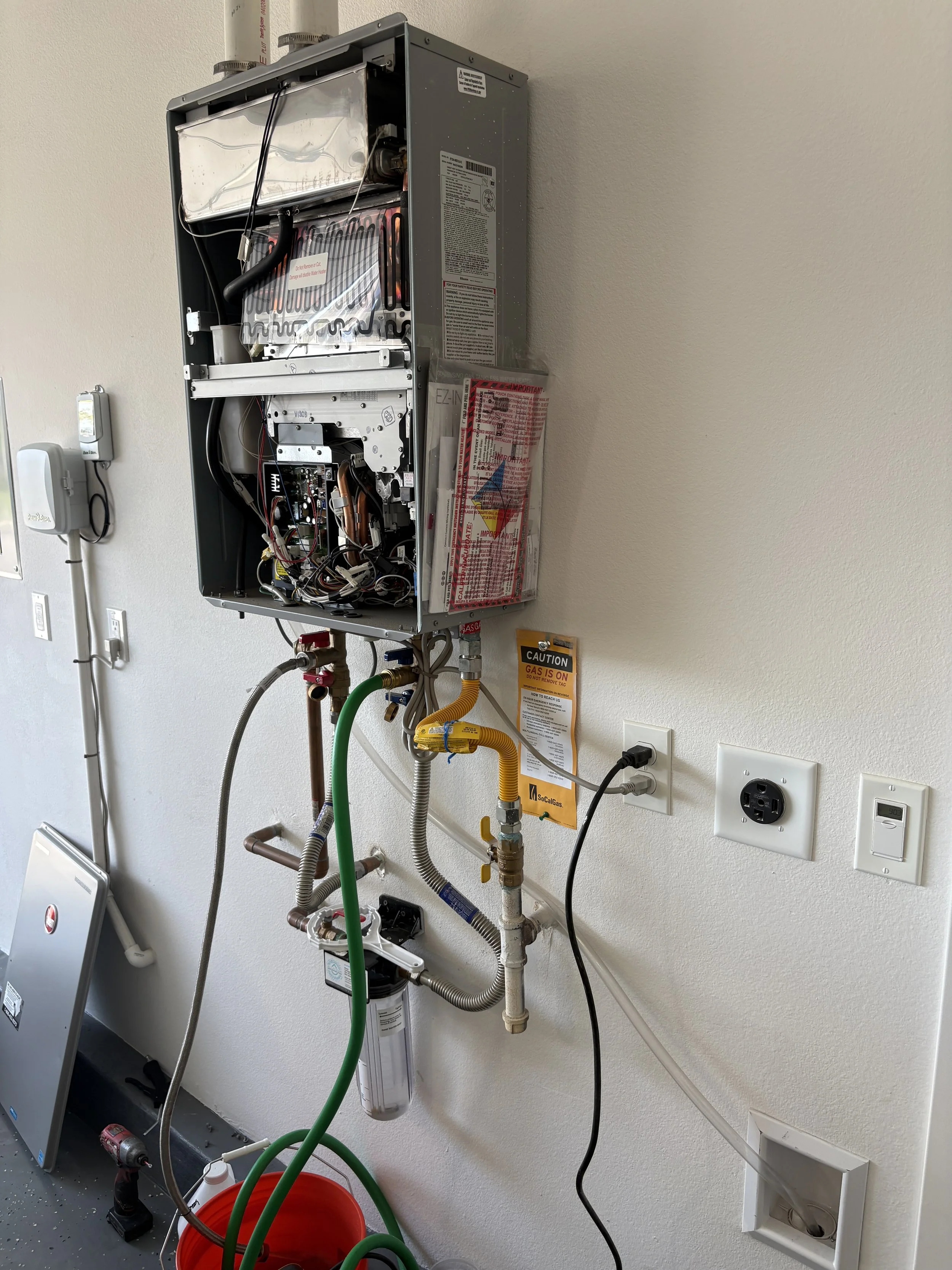 Open HVAC furnace or air conditioning unit mounted on a wall in a utility room, with connected pipes, wires, and a water filter below.