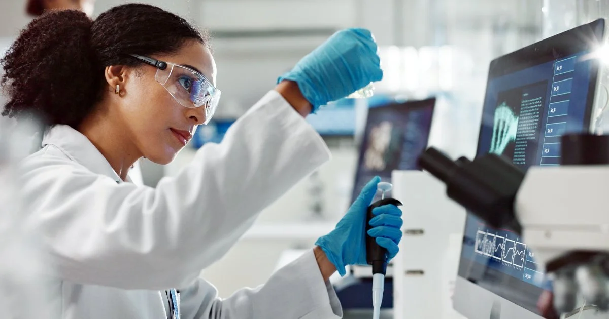 A scientist wearing safety glasses and blue gloves working in a laboratory, using a pipette, with medical images and data displayed on a computer screen.