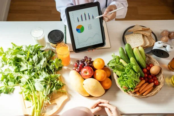 A person analyzing health data on a tablet with graph charts, surrounded by fresh fruits, vegetables, bread, and beverages on a kitchen table.