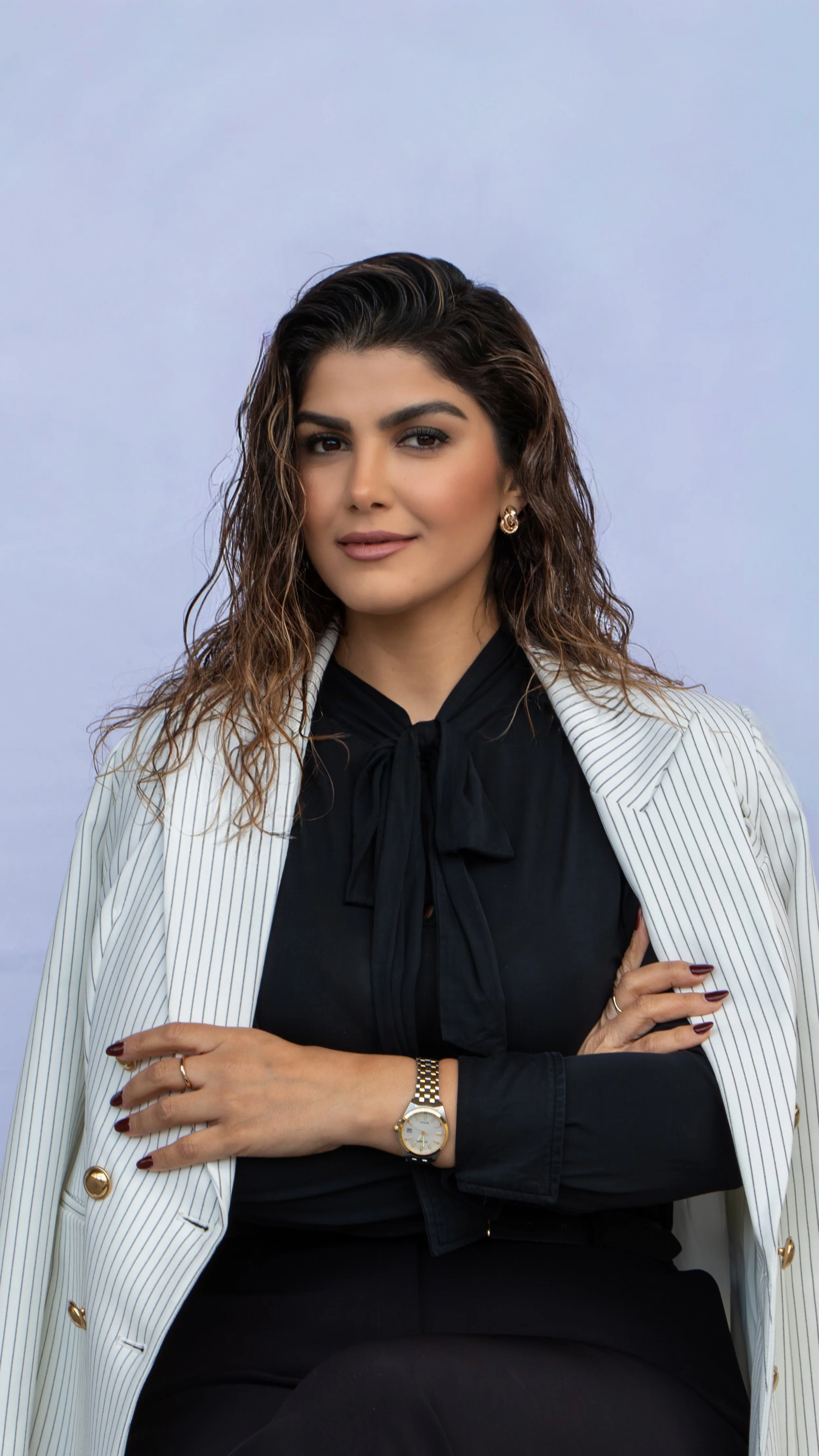 A confident woman with wavy hair, dressed in a black blouse with a tie neck and a white blazer with pinstripes, sitting against a light purple background.