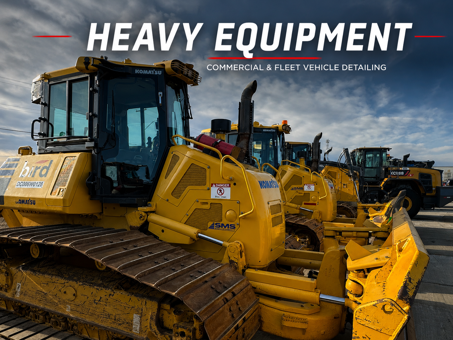 Line of yellow heavy construction equipment vehicles, including bulldozers and road graders, parked outdoors under a partly cloudy sky