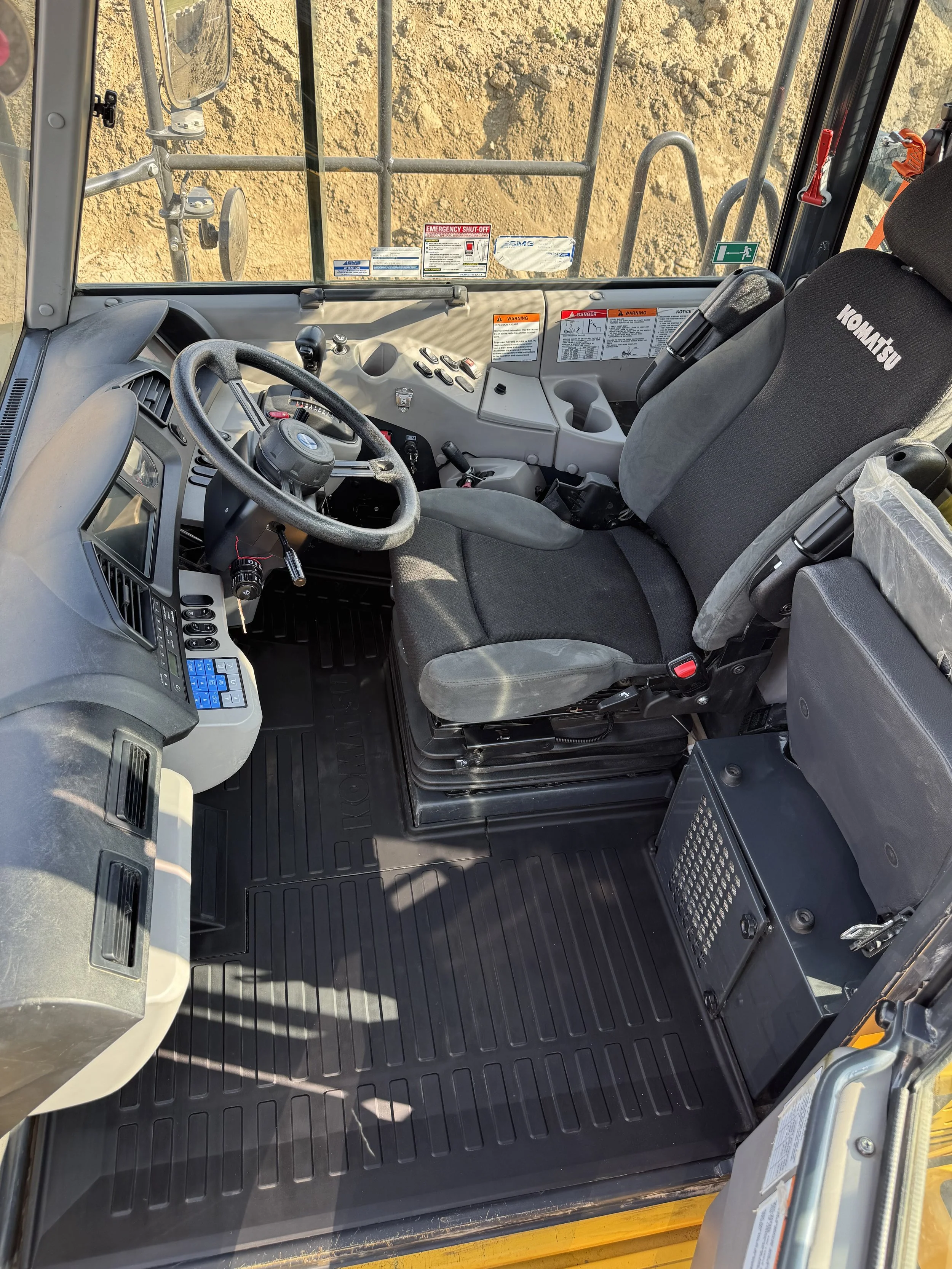 Interior view of construction equipment cab with steering wheel, control panels, gray seat, and safety stickers, with a construction site visible through the window.