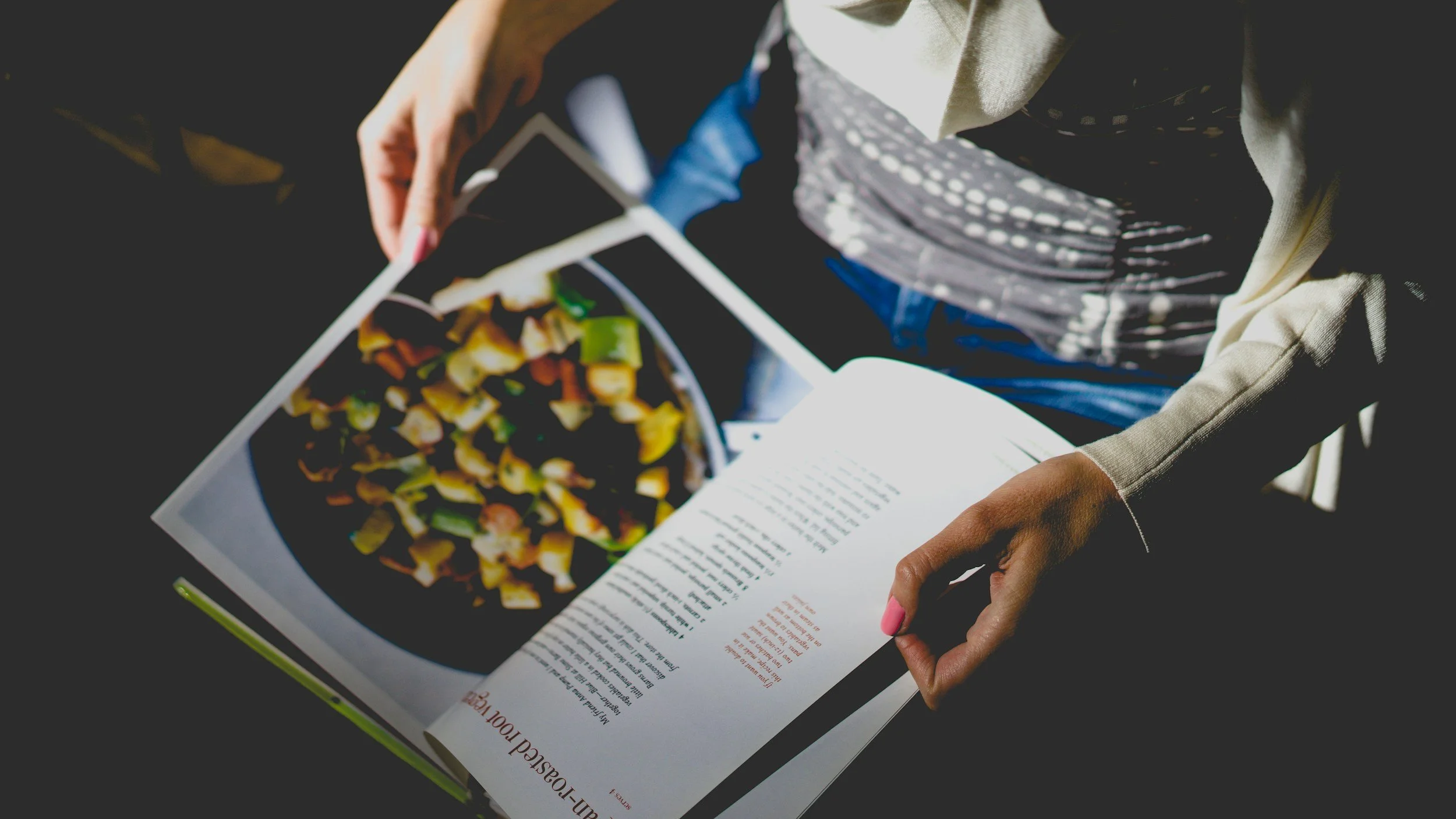 Person holding a magazine open to a food recipe page, with a colorful image of a vegetable dish.