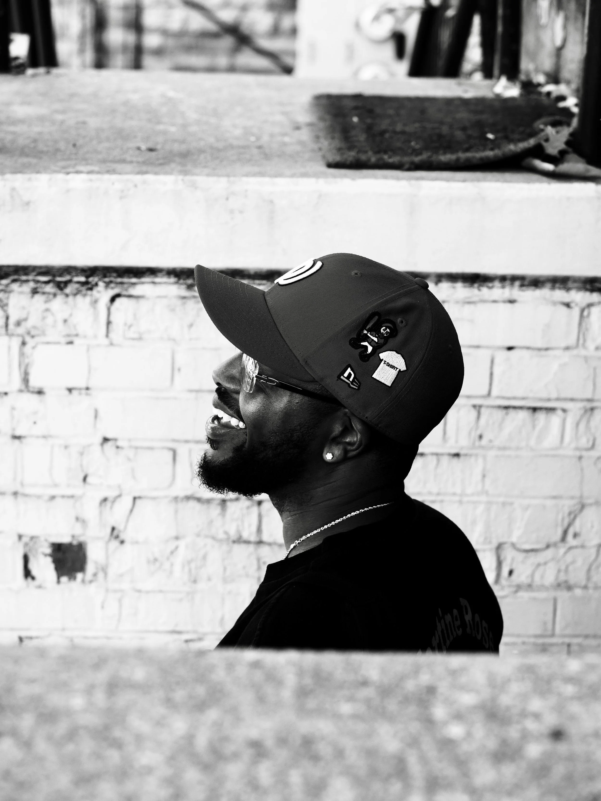 A black and white photo of a man smiling and looking to the left, wearing a baseball cap and glasses, with a brick wall in the background.