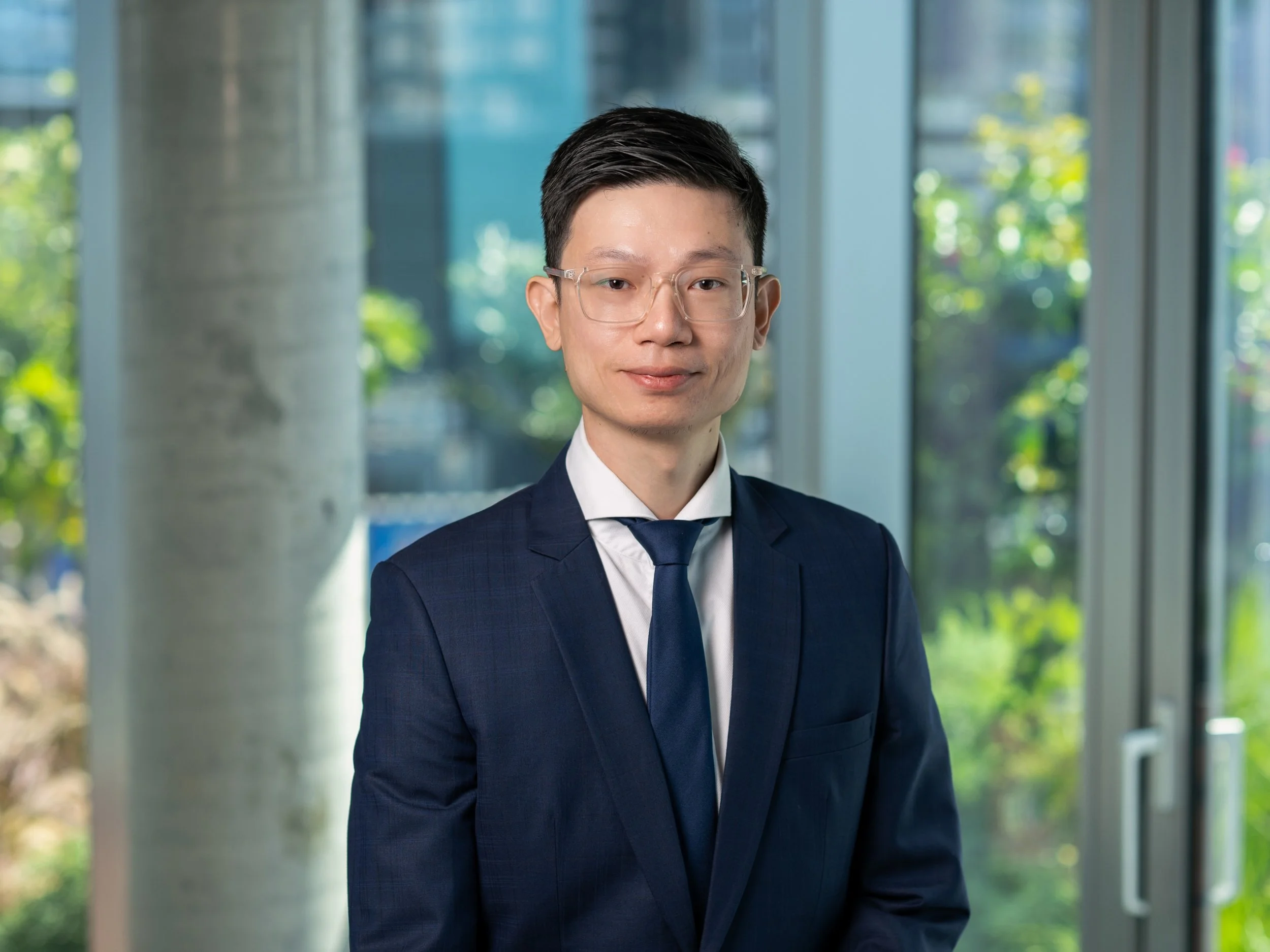 A professional man in a dark blue suit, white shirt, and matching blue tie standing indoors near large glass windows with greenery outside, smiling slightly.
