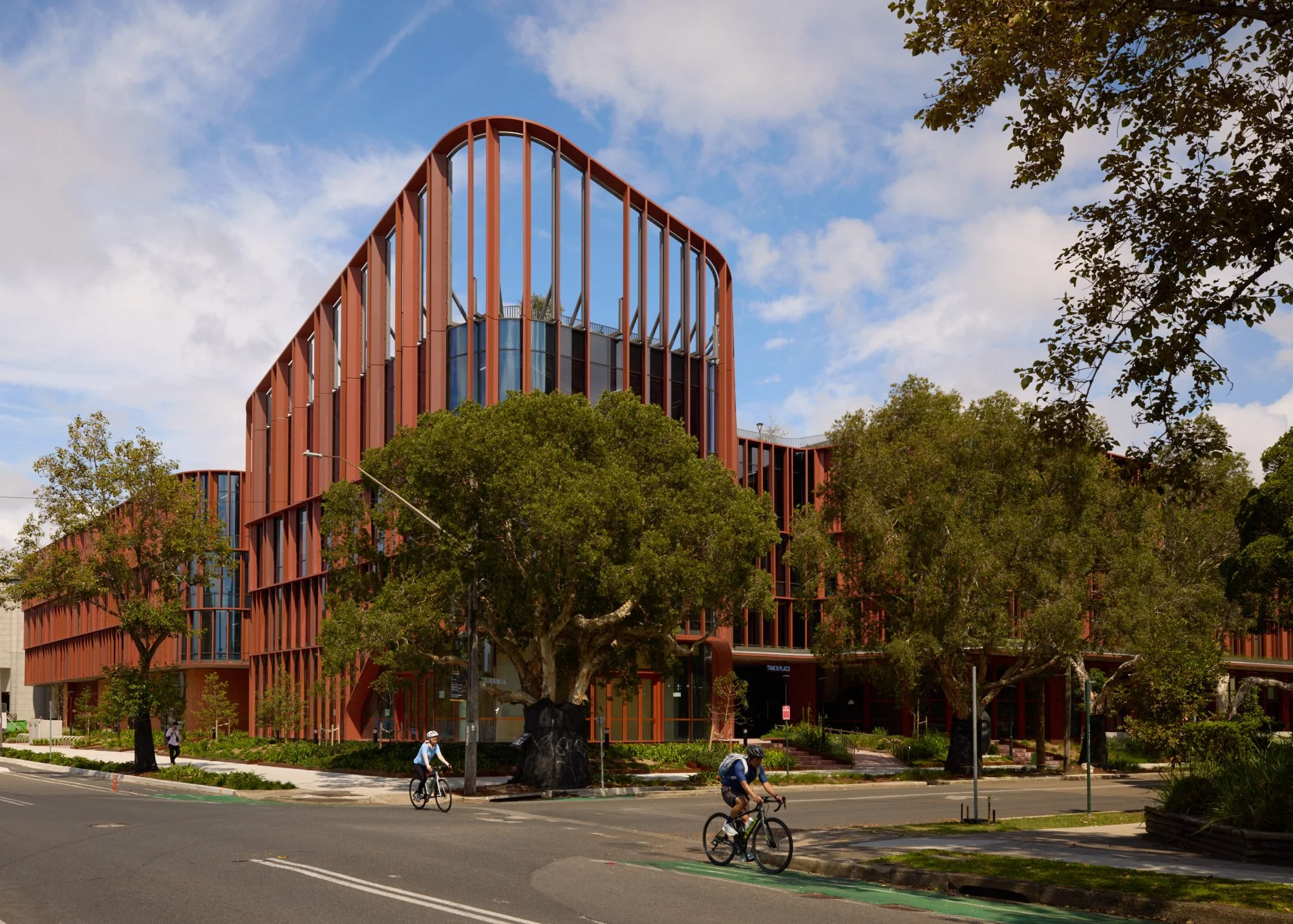 Modern multi-story building with red vertical beams and large windows, surrounded by green trees and people biking on the street under a partly cloudy sky.