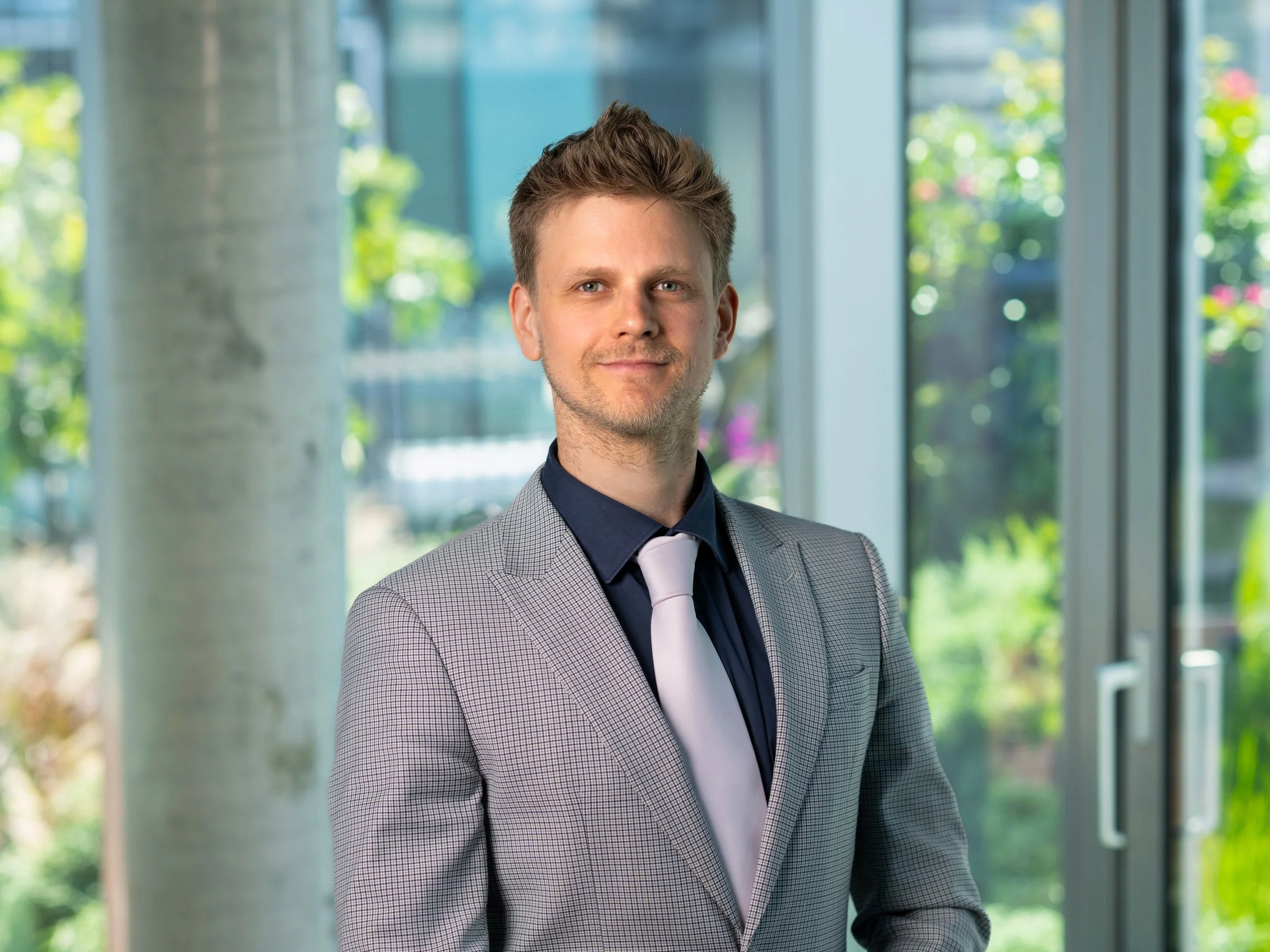 A man in a gray suit with a dark shirt and light-colored tie standing in front of large glass windows showing trees and greenery outside.
