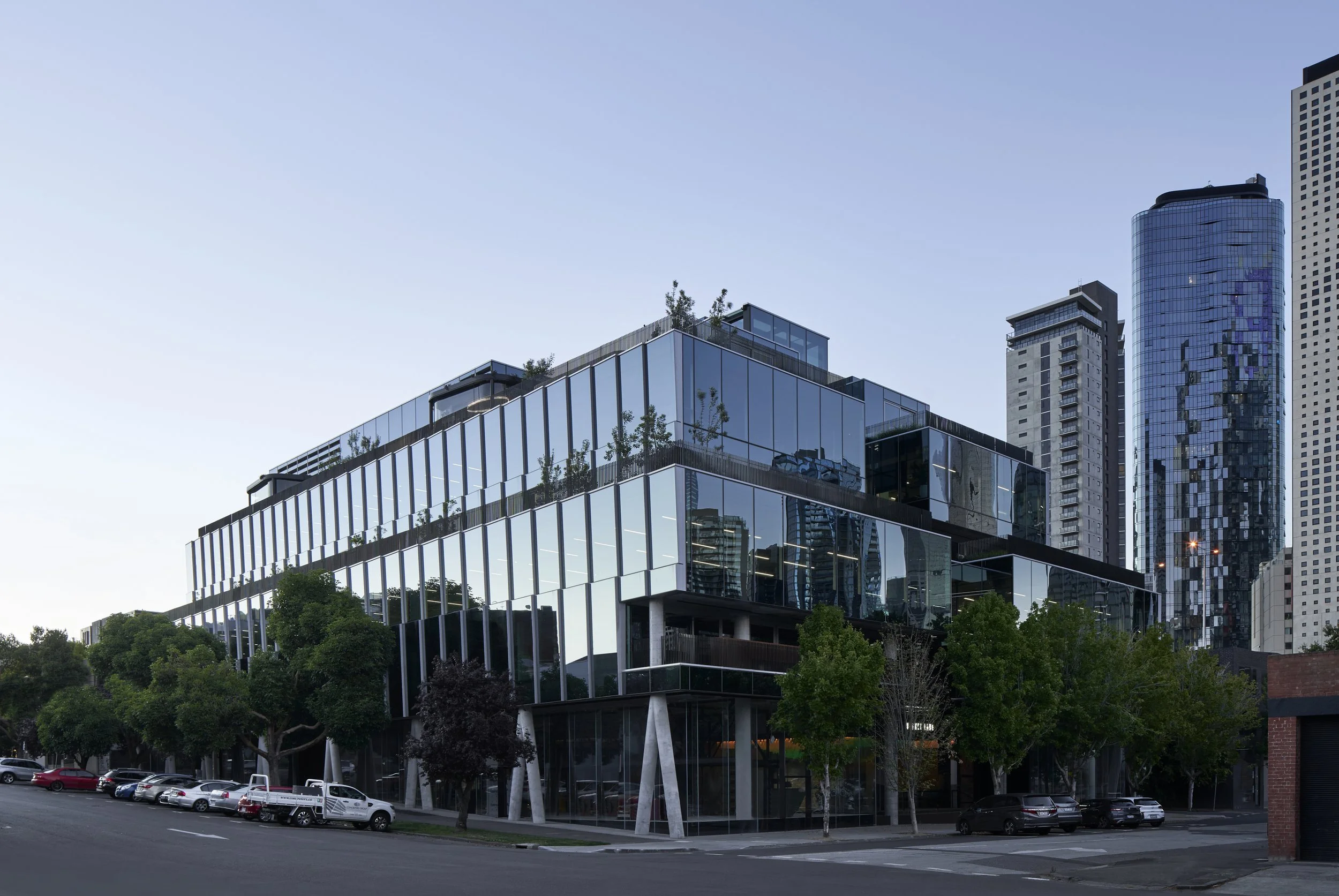 Modern multi-story office building with glass facade, reflecting city skyscrapers, surrounded by trees and parked cars.