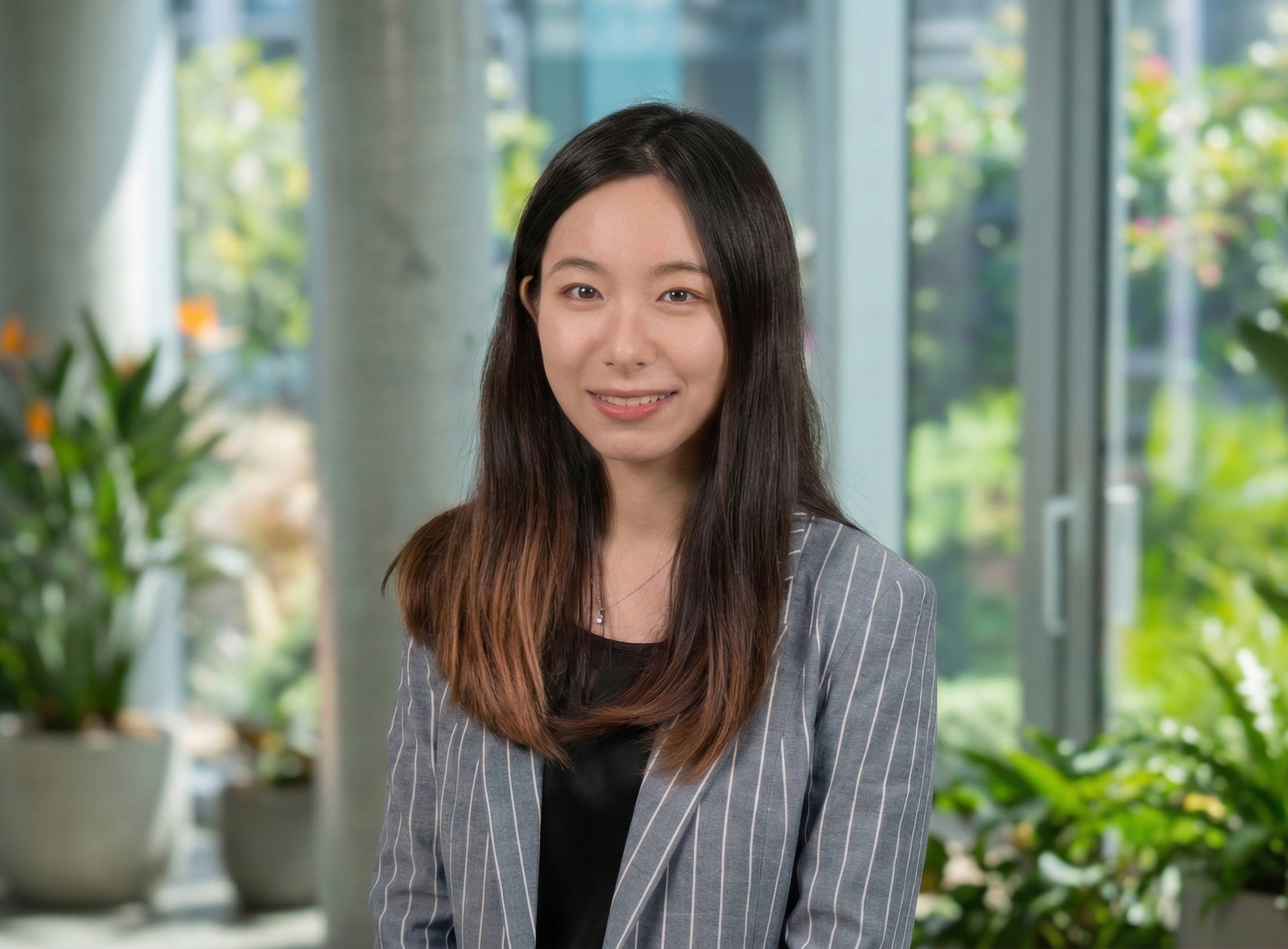 A young woman with long dark hair, wearing a gray pinstripe blazer, smiling at the camera with a cityscape background.