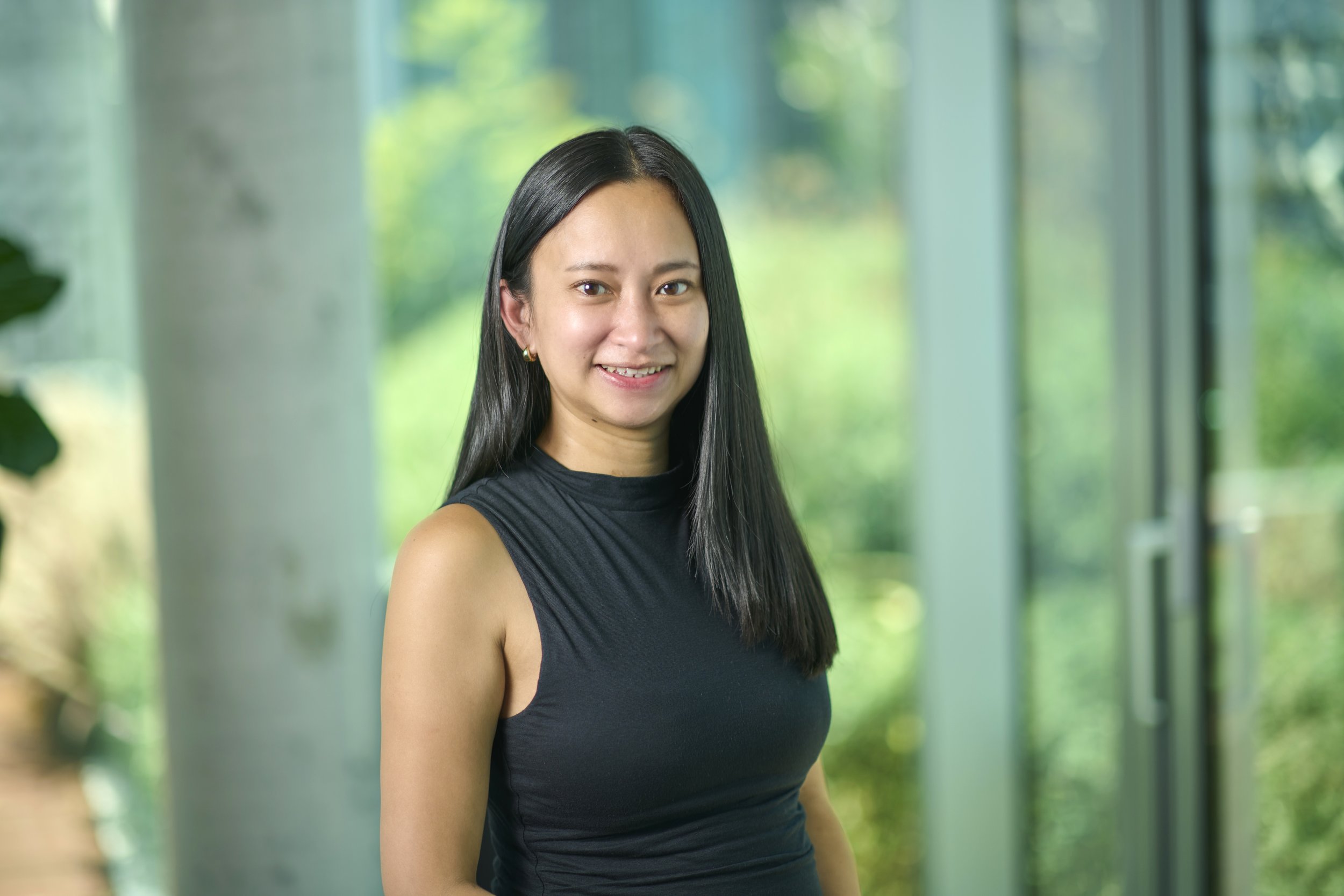 A woman with long black hair wearing a sleeveless black top, standing indoors with large windows and greenery outside.