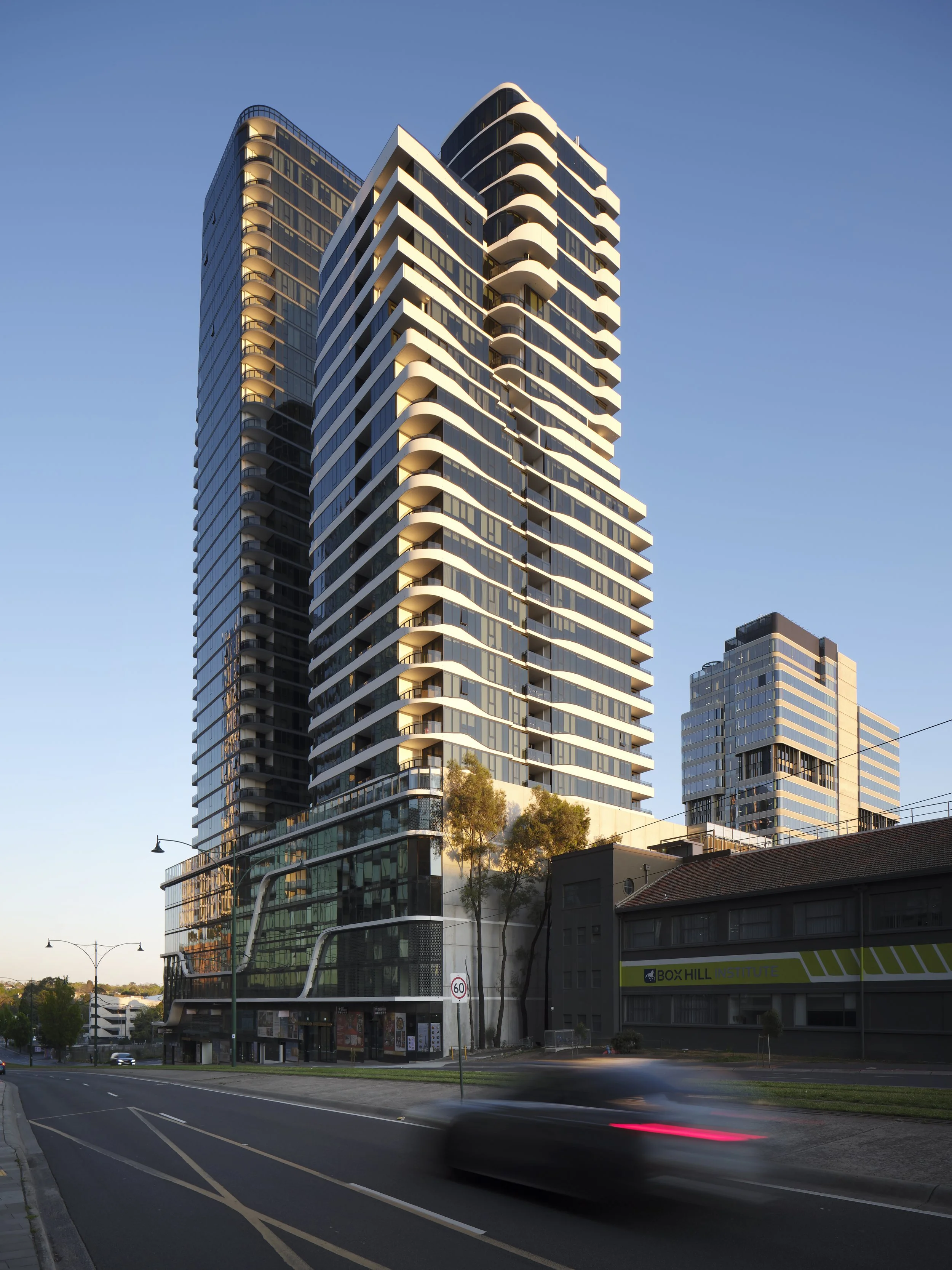 A modern high-rise building with curved balconies and glass windows, situated along a city street at dusk.