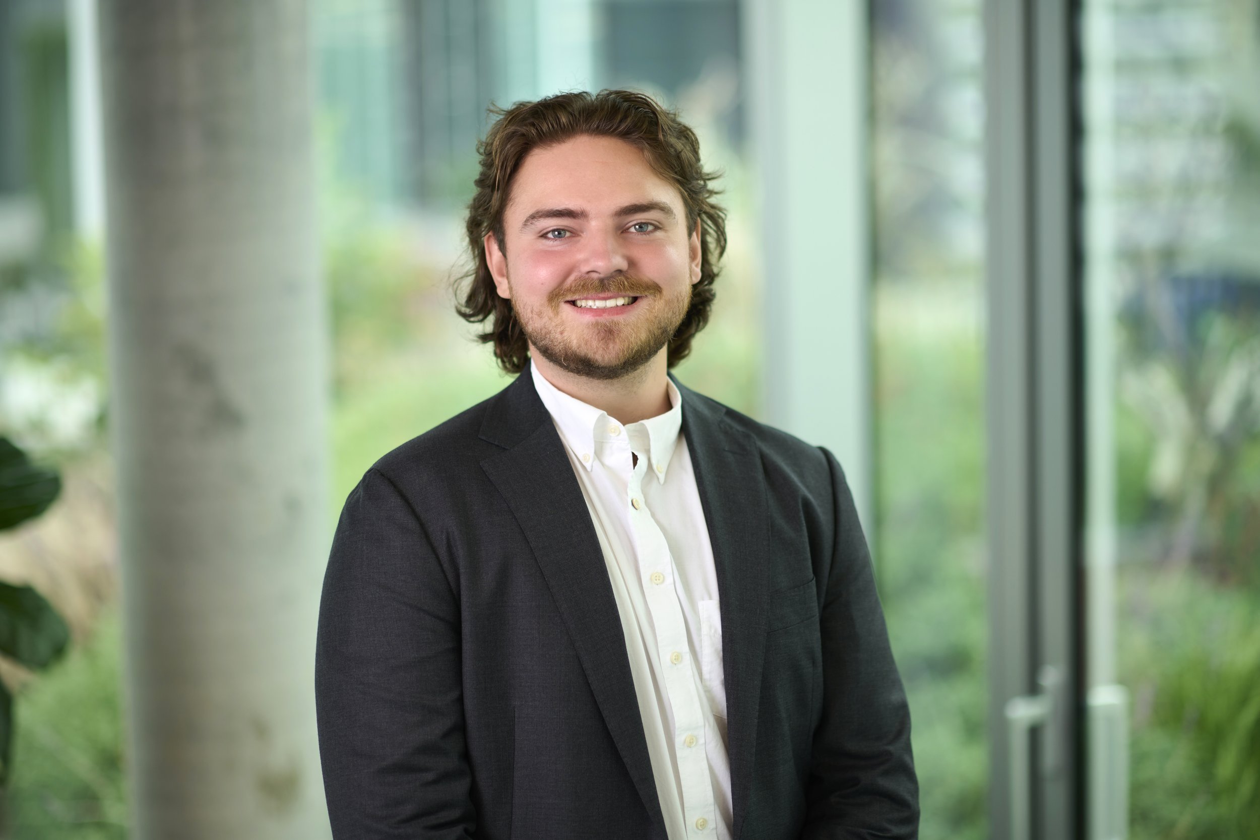 A smiling man wearing a dark blazer and white shirt, standing indoors with large windows showing greenery outside.