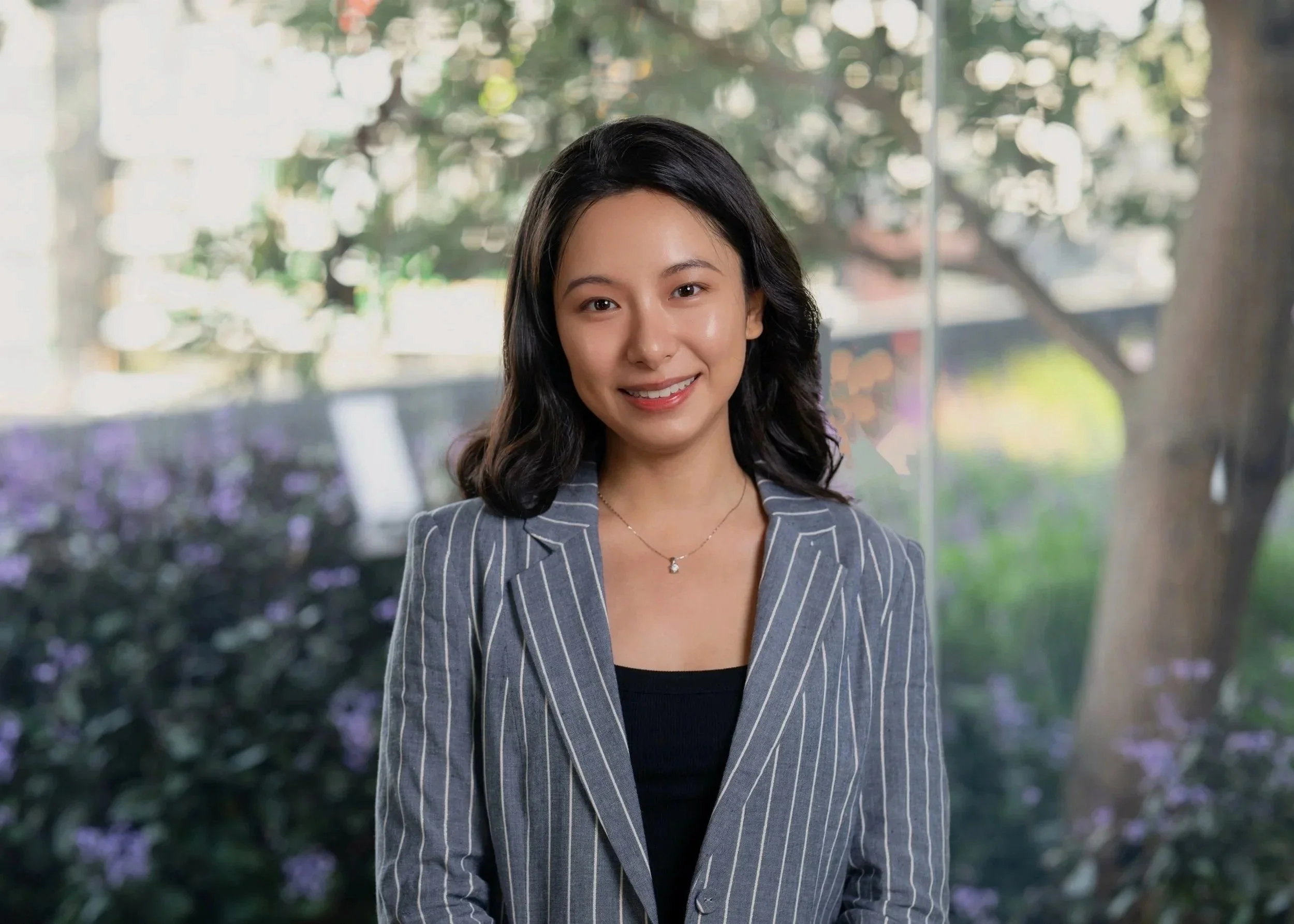 A young woman with long dark hair, wearing a gray pinstripe blazer, smiling at the camera with a cityscape background.