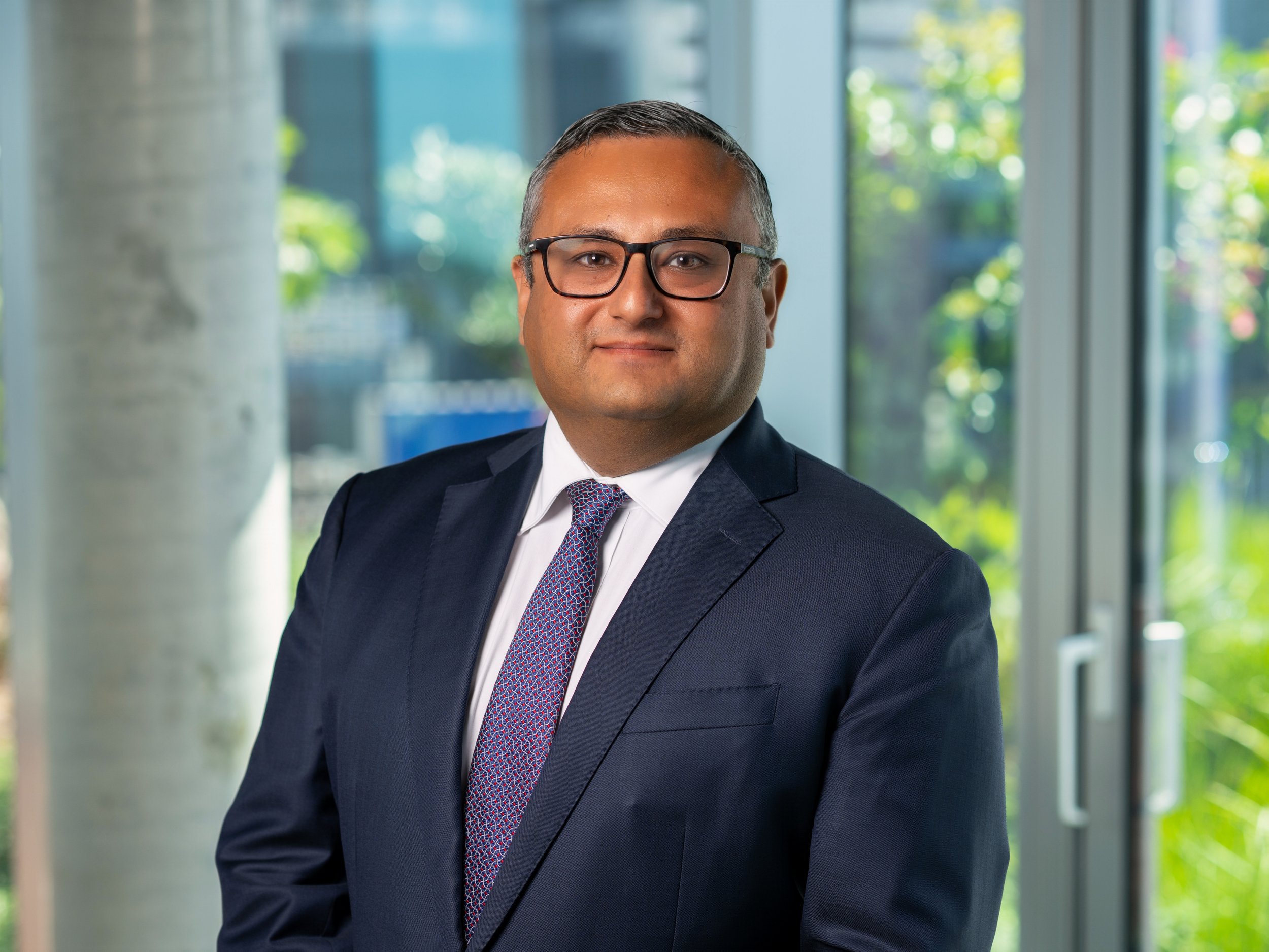 A professional man with glasses in a business suit standing indoors with a glass door and greenery outside.