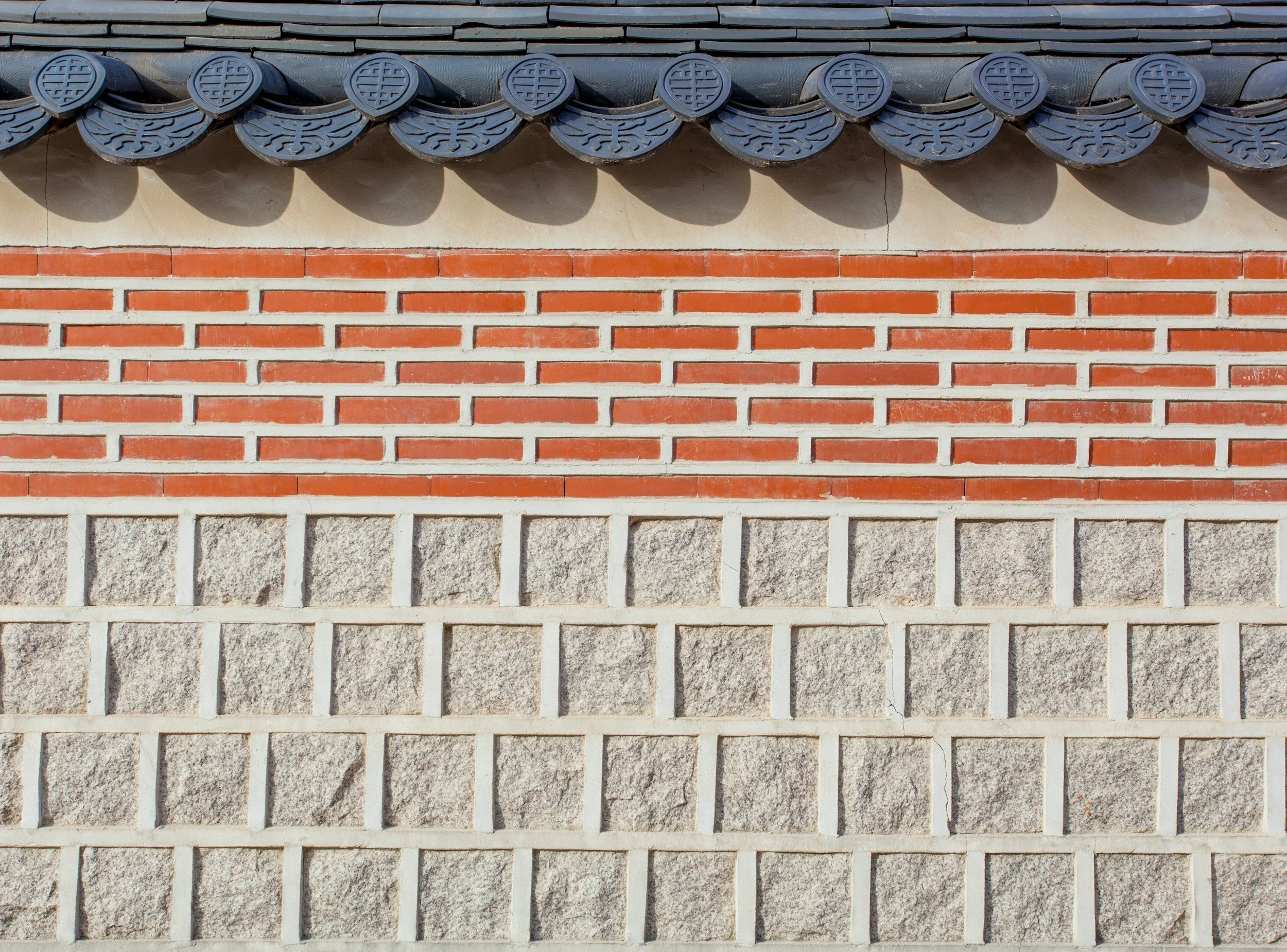 Close-up of traditional Asian-style tiled roof with ornate dark gray roof tiles, red brick wall in the middle, and gray stone blocks at the bottom.