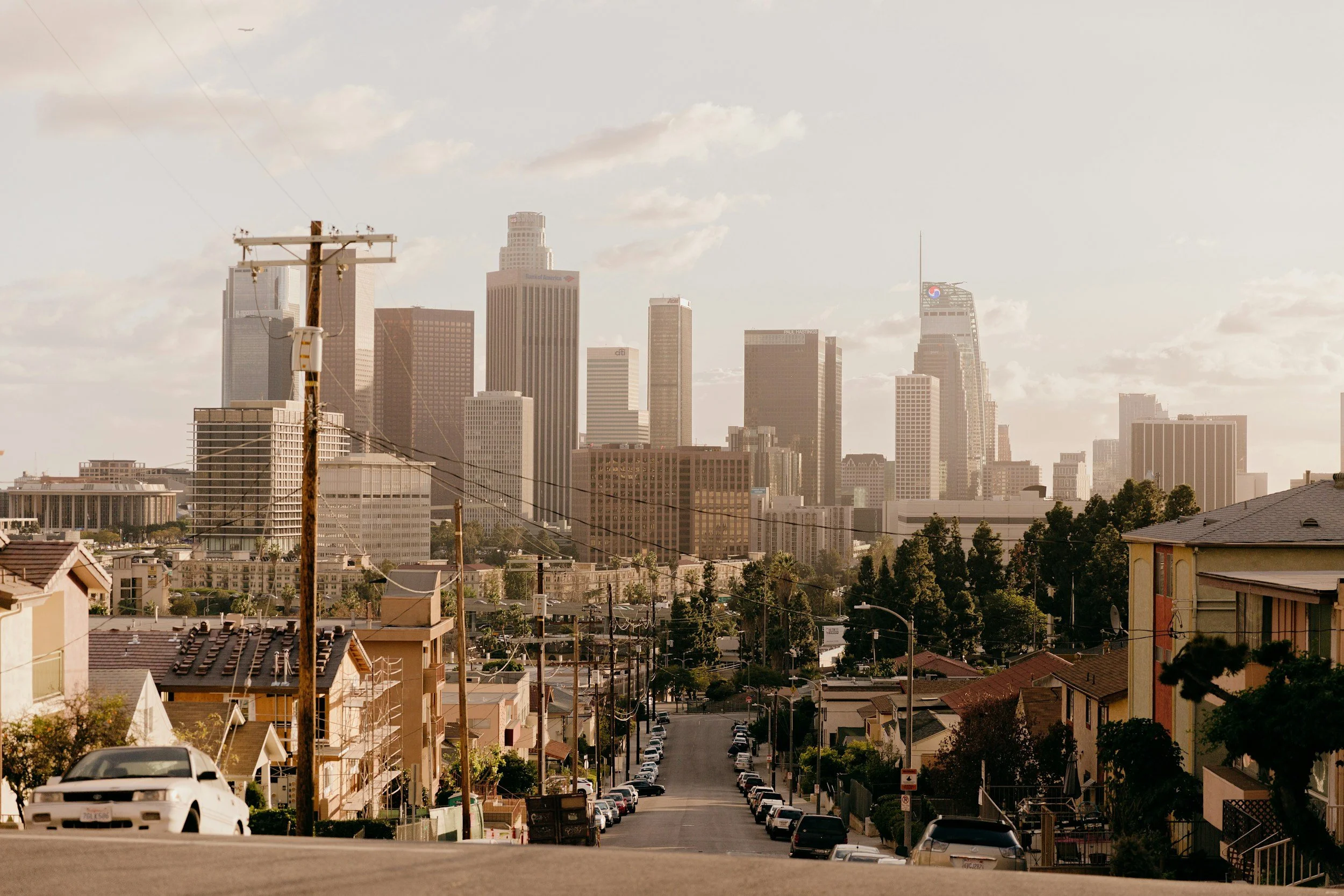 A city skyline with tall skyscrapers viewed from a residential neighborhood street with houses, parked cars, and utility poles.