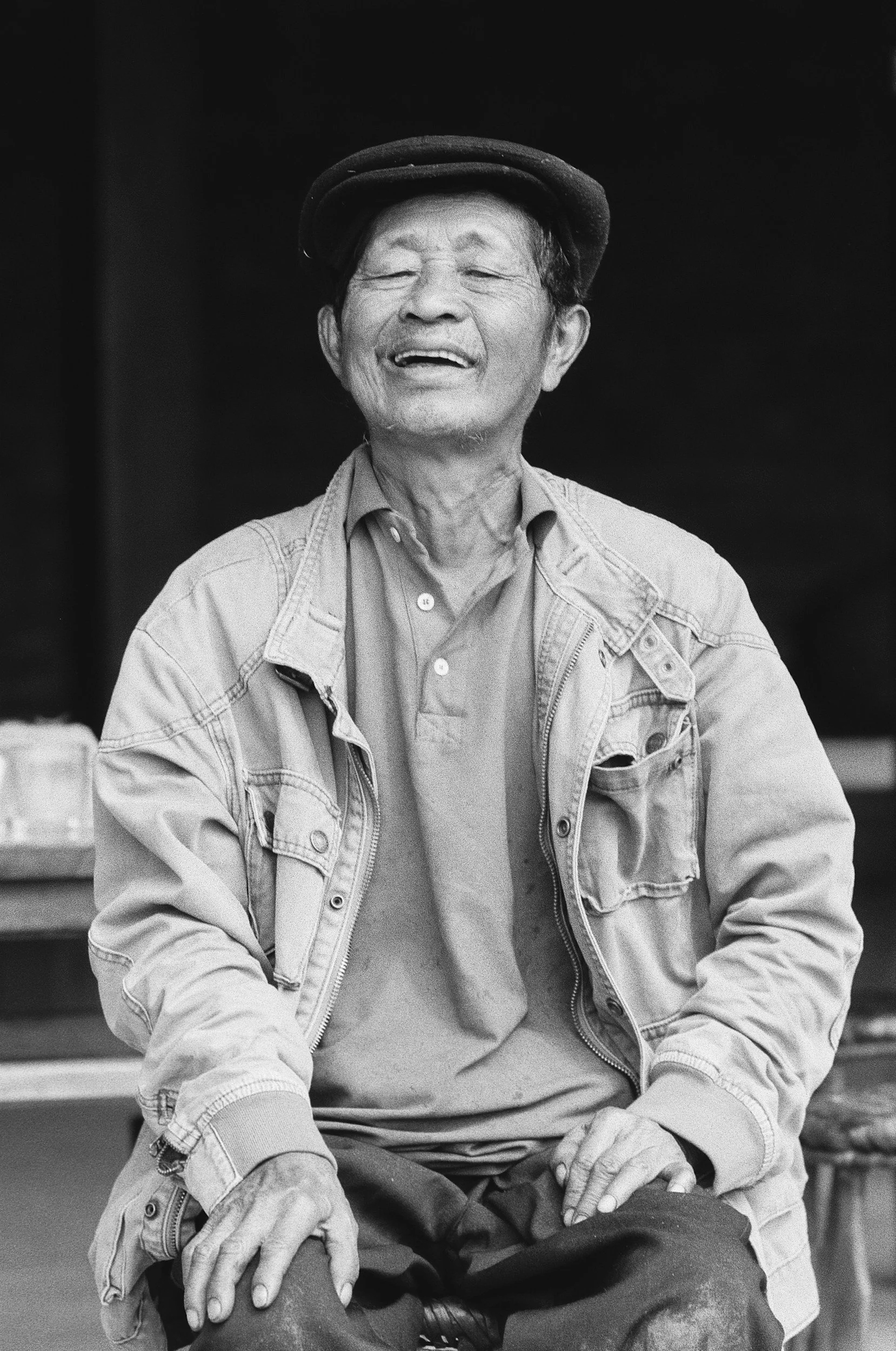 Black-and-white photo of an elderly man smiling, wearing a cap, a denim/jacket over a button-up shirt, and seated with his hands on his lap.