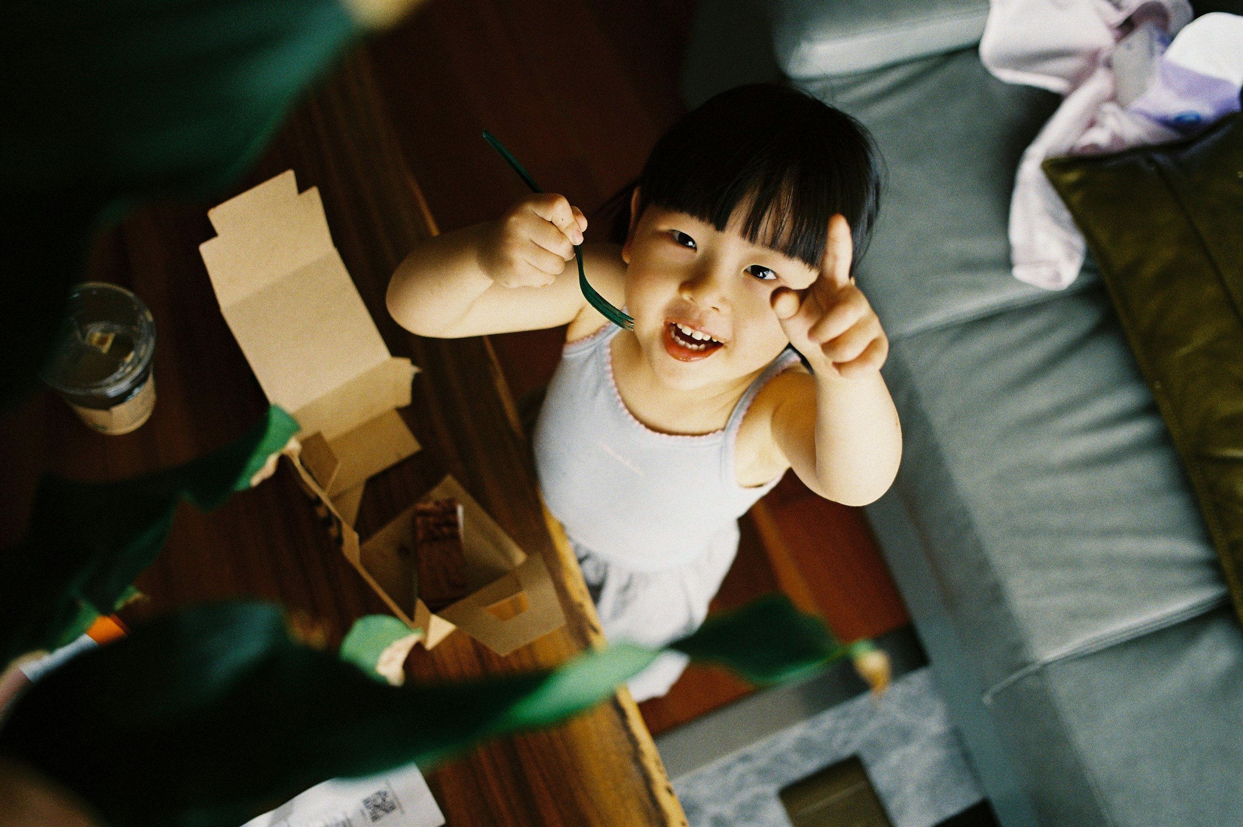 A young Asian girl with black hair and bangs, wearing a white sleeveless top, smiling and pointing at the camera while holding a fork, standing next to an open cardboard takeout box on a wooden table.