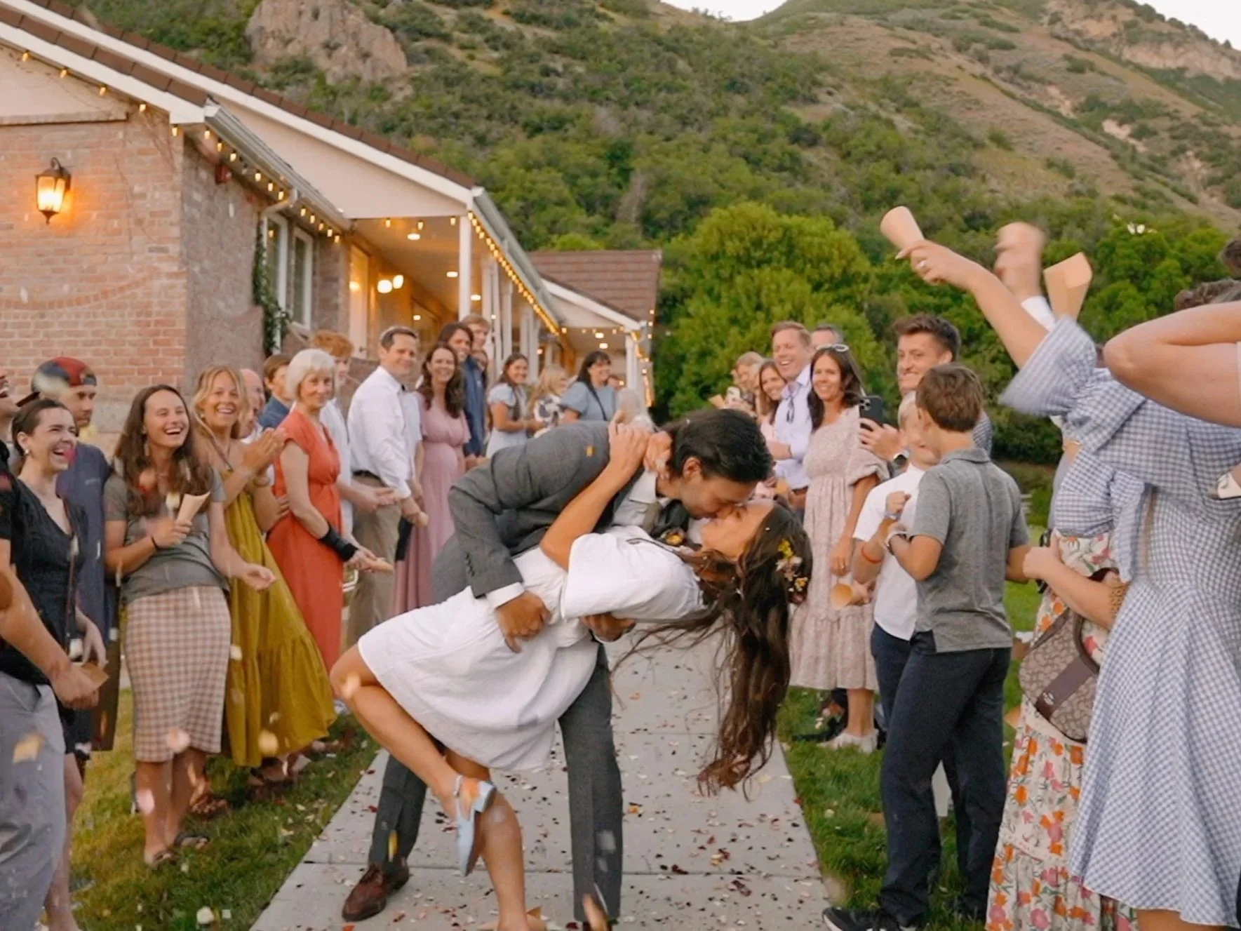 A wedding celebration with a bride and groom kissing as guests cheer and throw flower petals outside a rustic building with string lights, mountains, and greenery in the background.