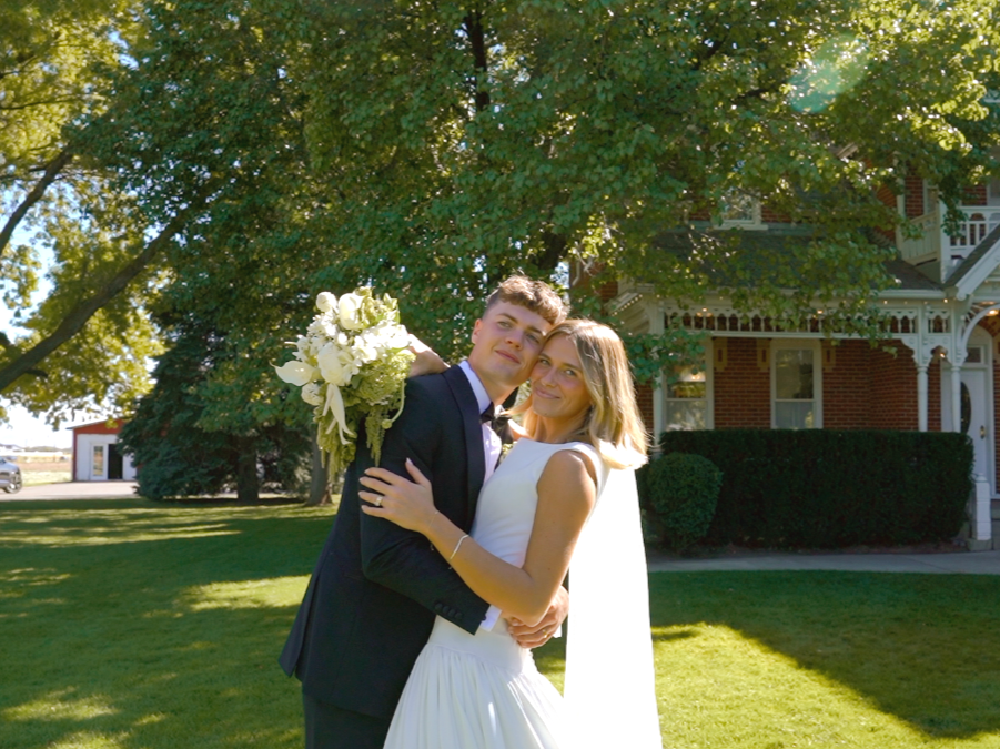 A newlywed couple in wedding attire embracing outdoors in front of a large tree and a Victorian-style house, with sunlight filtering through the branches.