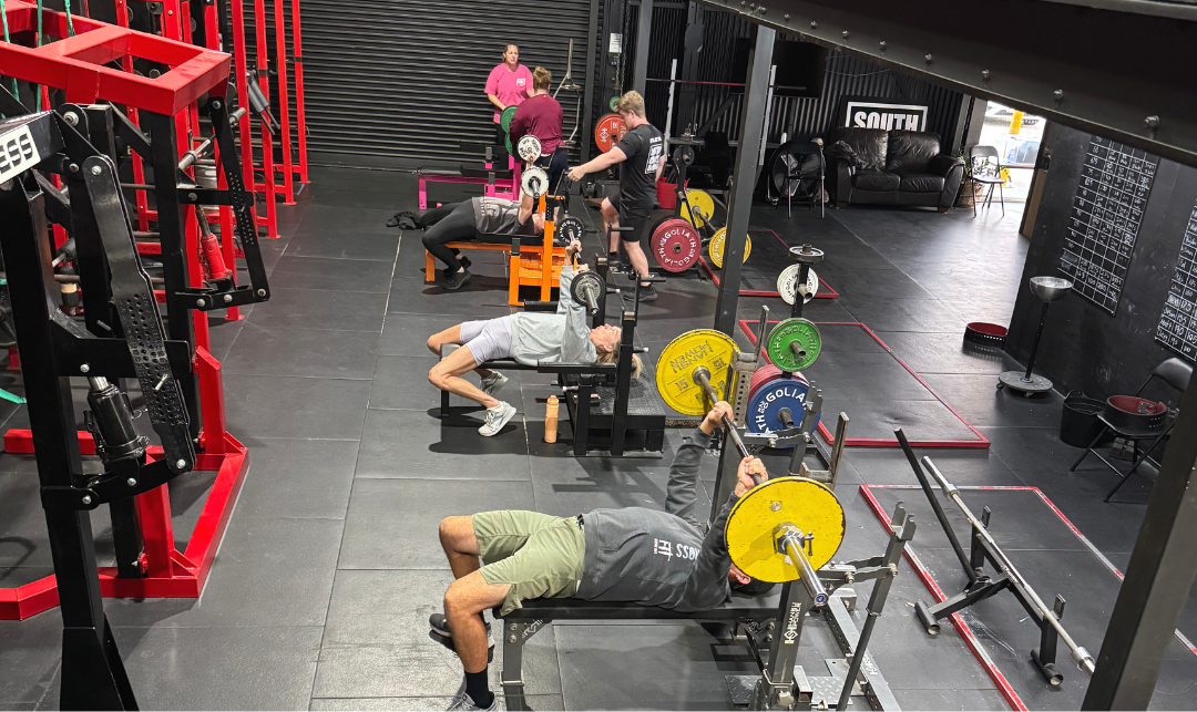 People lifting weights and doing exercises at a gym with equipment such as barbells, benches, and squat racks.