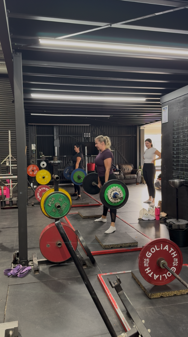 Women lifting weights in a gym with weight plates and barbells, with fitness equipment and black walls in the background.