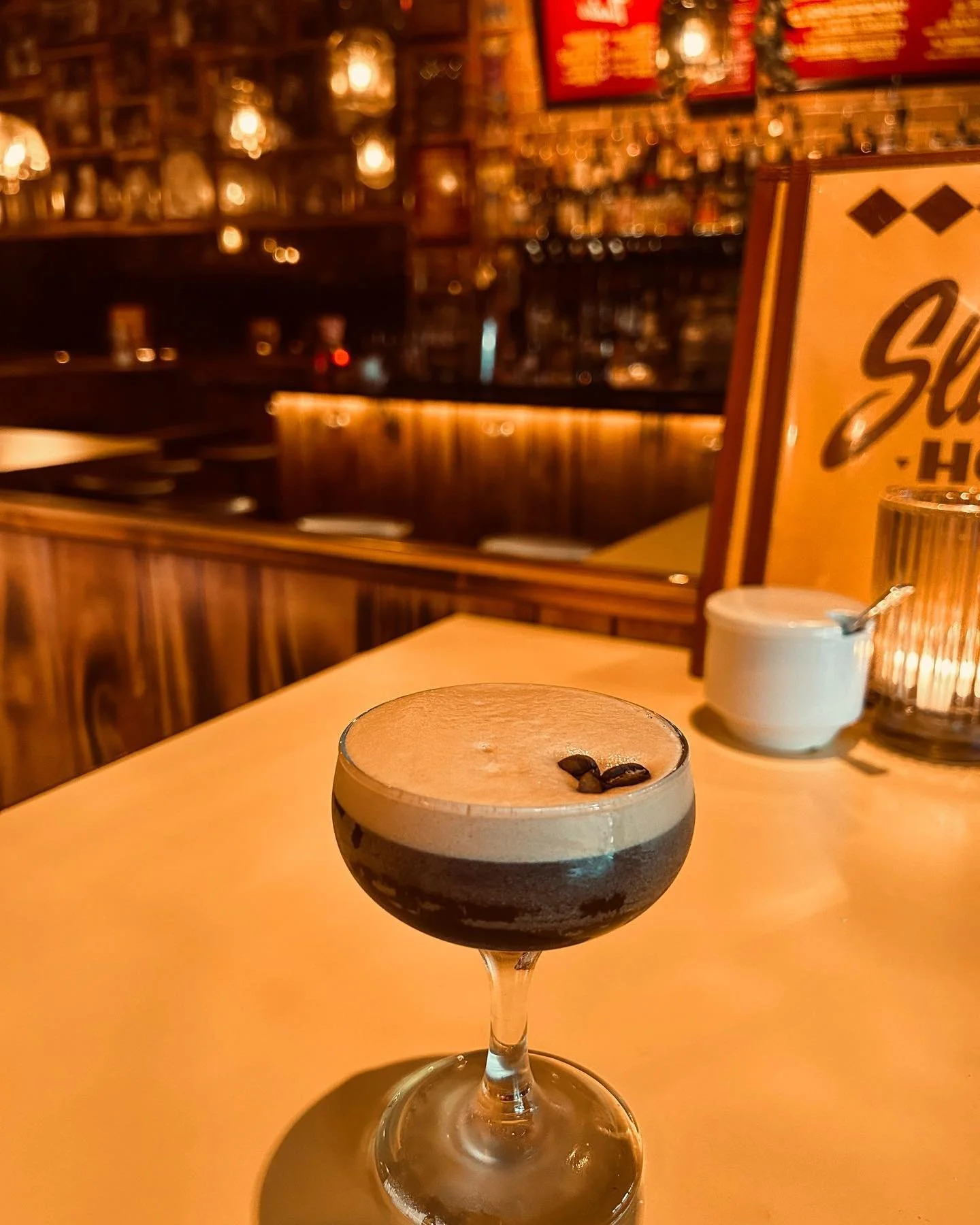An espresso martini with a creamy top and coffee beans garnishing it is on a table in a dimly lit restaurant or bar with warm lighting and brick walls.