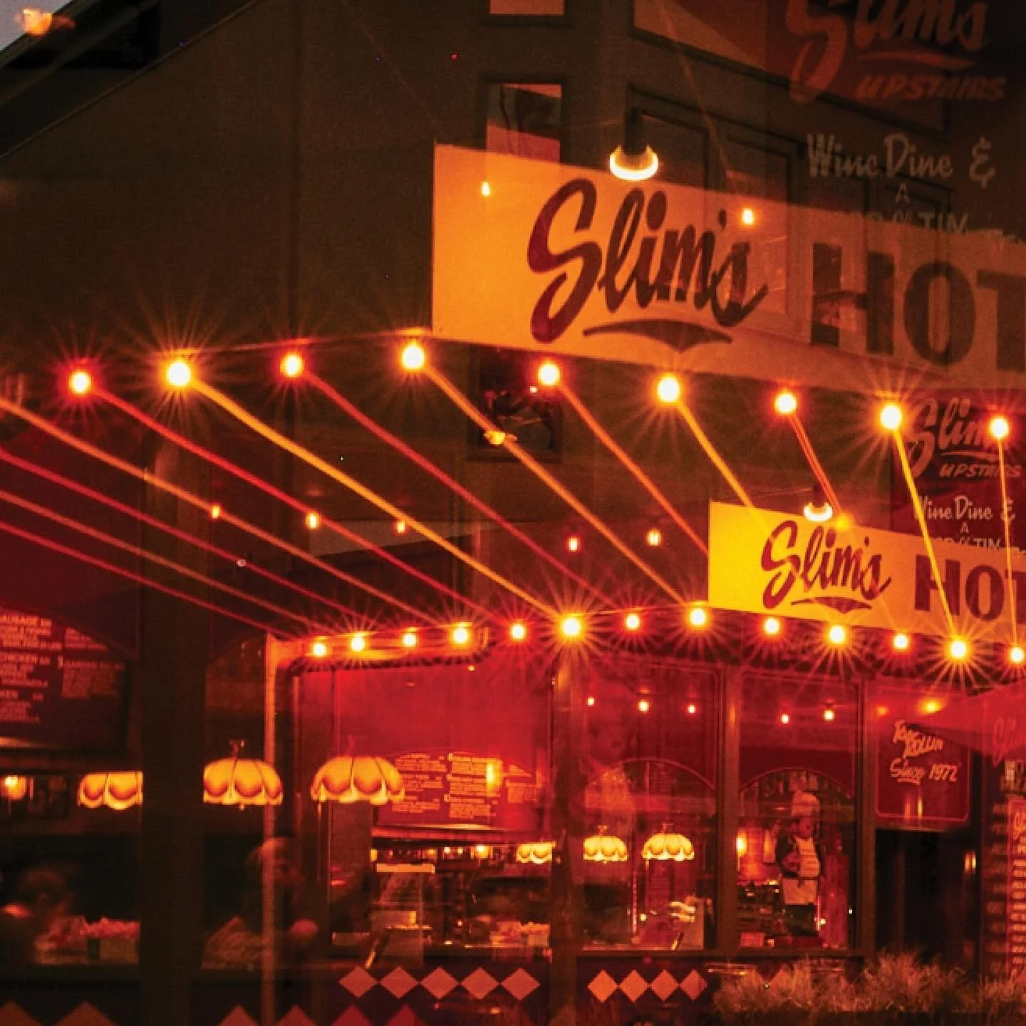 Night view of Slim's restaurant with illuminated red string lights and warm yellow hanging lamps inside, reflected on the glass window.