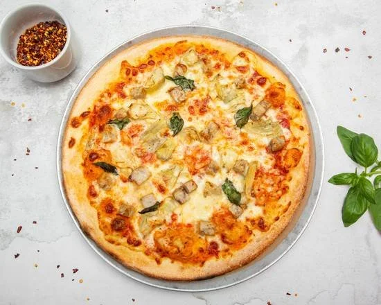 Cheese pizza topped with basil and chicken slices on a round metal tray, with a small cup of red pepper flakes and fresh basil leaves nearby.