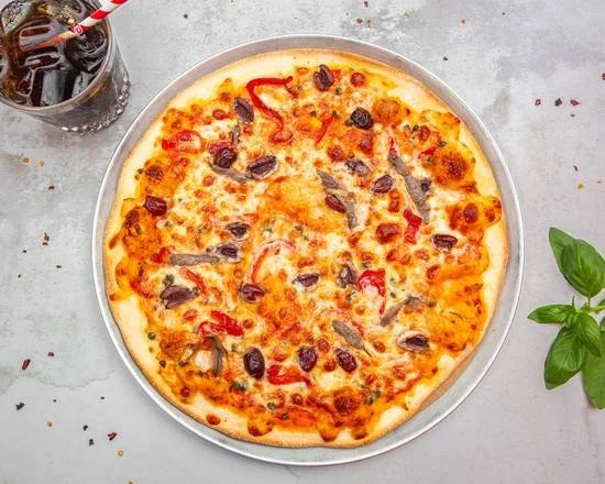 A pizza with cheese, black olives, red peppers, and possibly sausage slices, on a round metal tray, with a glass of soda and basil leaves nearby on a gray surface.