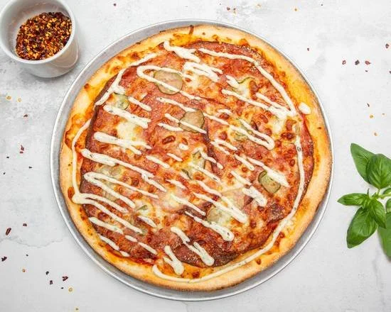 Whole pizza topped with melted cheese, tomato sauce, and jalapenos, drizzled with white sauce, on a metal tray. A small cup of red pepper flakes and fresh basil leaves are nearby.