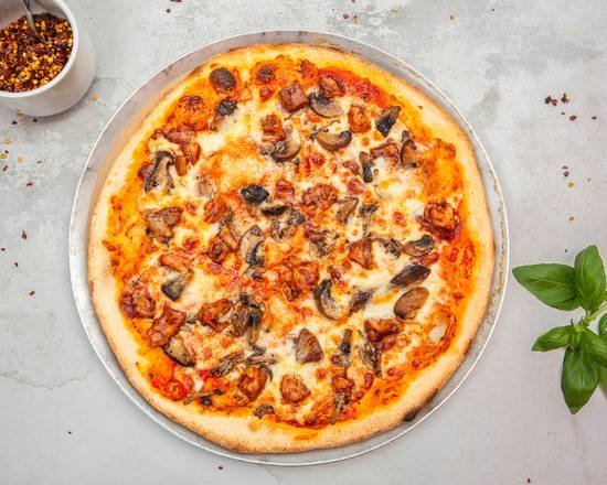 A cheese pizza topped with mushrooms and pepperoni on a round tray, with a bowl of red chili flakes and a small plant nearby on a gray surface.
