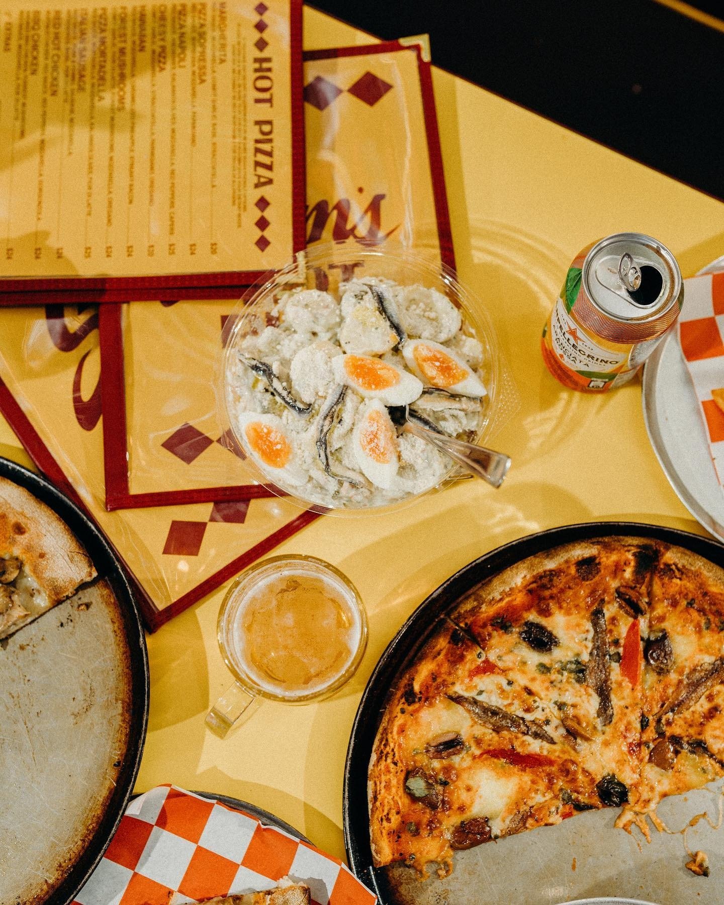Pizza slices, a bowl of salad with eggs, a can of soda, a glass of beer, and the paper menus on a yellow table in a pizzeria.