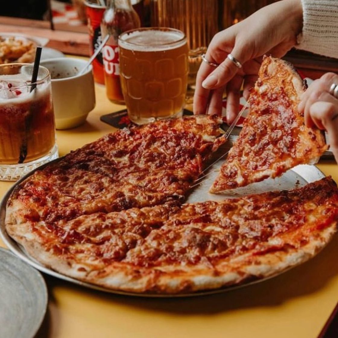 Person taking a slice of large pepperoni pizza at a restaurant table with two glasses of beer and other drinks visible.