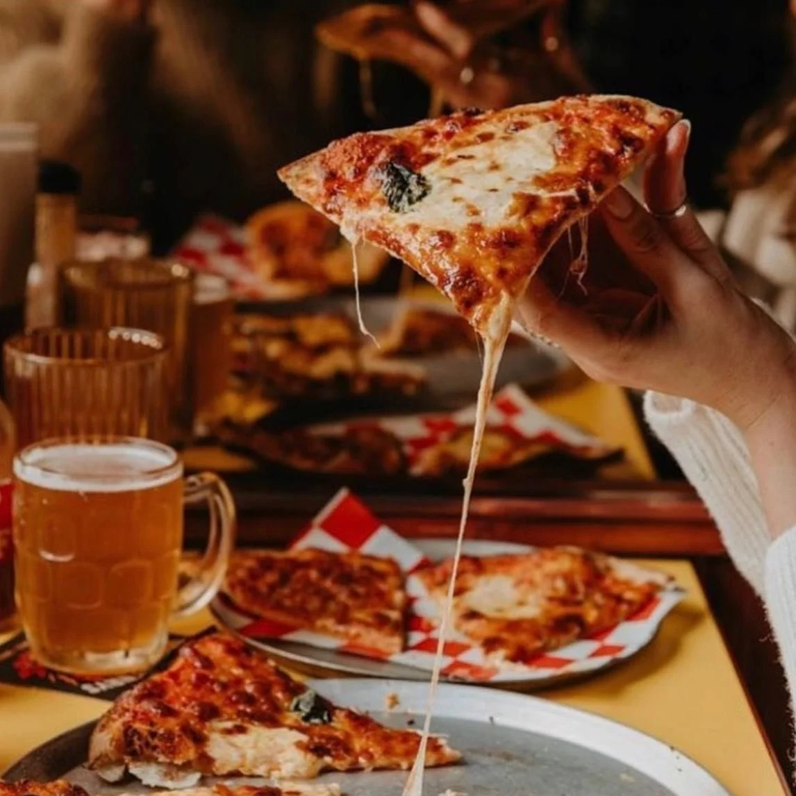 Person holding a slice of cheese pizza with melted cheese stretching, with beer mugs and pizza in the background at a pizza restaurant.