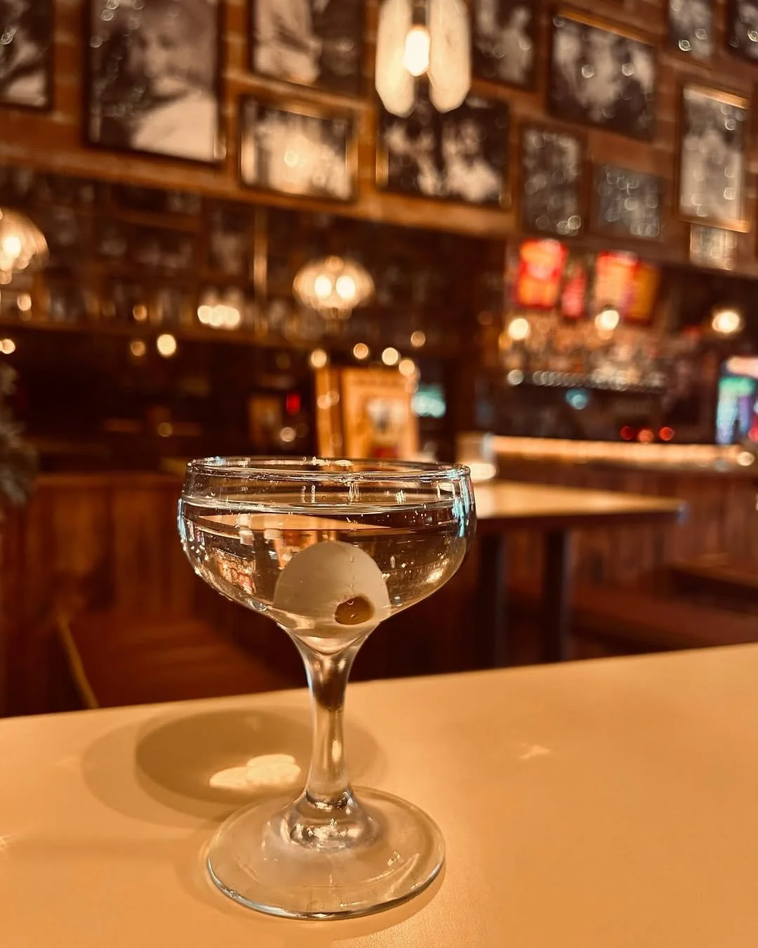 A cocktail glass with an olive, filled with a clear drink, on a table in a dimly lit bar or restaurant with wooden decor and framed pictures on the wall.