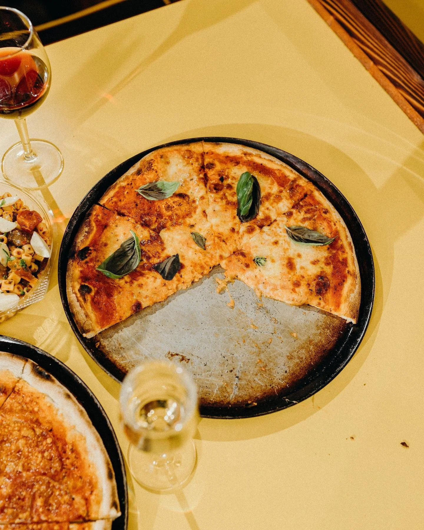 A partially eaten cheese pizza with basil garnish on a black pizza pan, a glass of red wine, a pasta salad, and a glass of white wine on a yellow table.