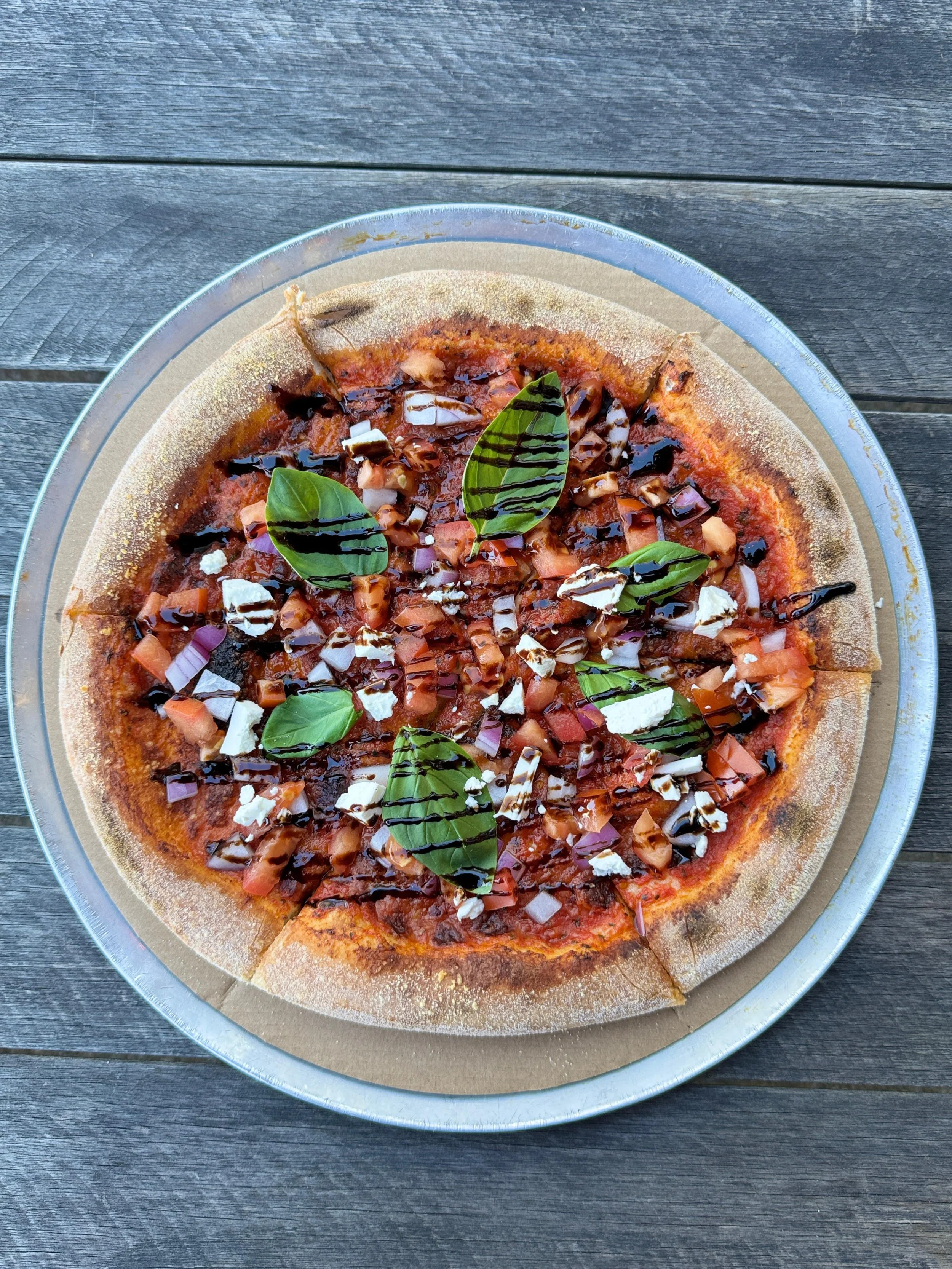 A pizza with toppings including chopped onions, diced tomatoes, and basil leaves, drizzled with balsamic glaze, on a metal tray and placed on a dark wooden surface.