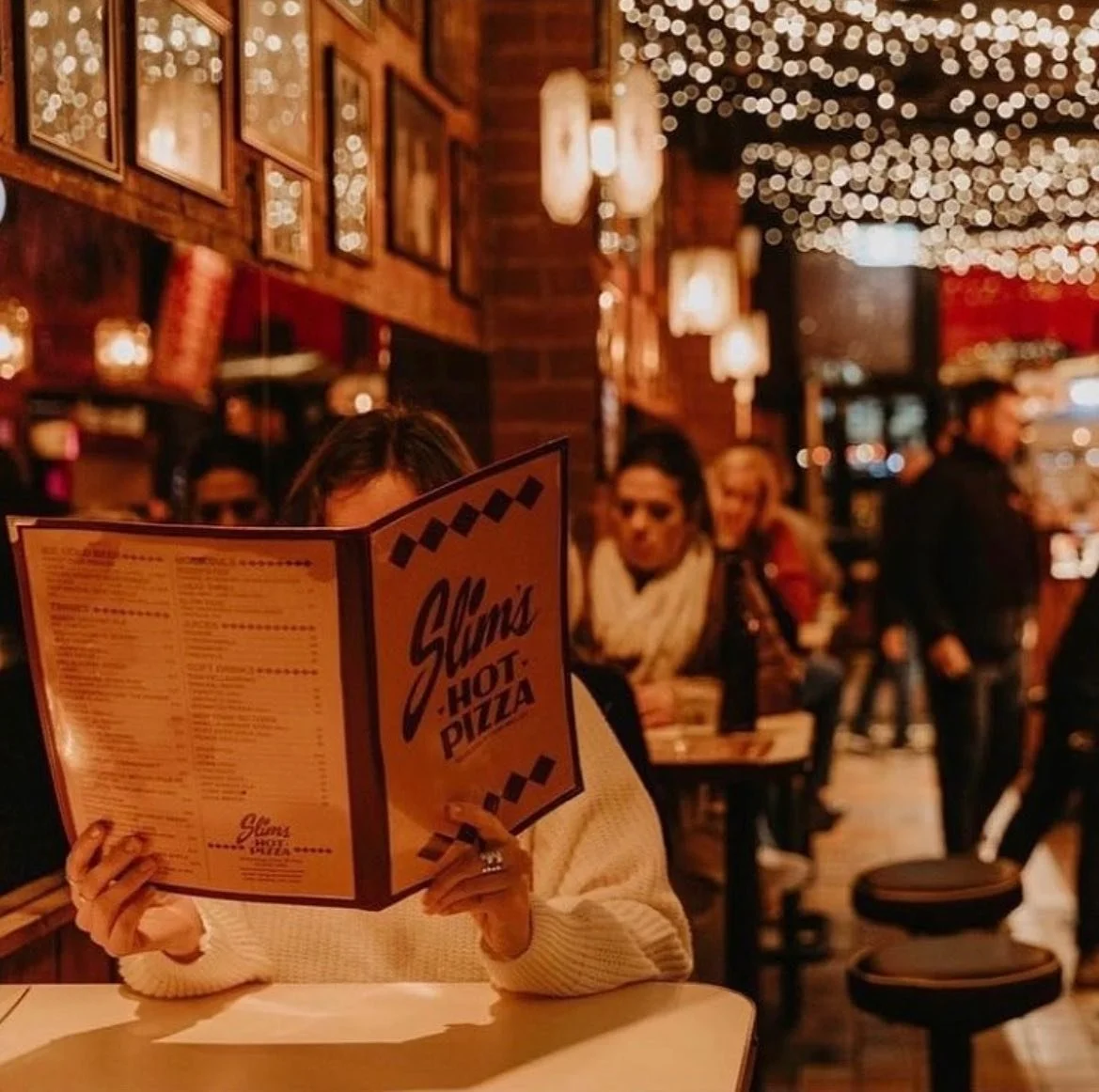 A woman in a white sweater sitting at a table in a dimly lit restaurant, reading a menu titled "Slims Hot Pizza" with other patrons in the background and string lights overhead.