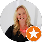A smiling blonde woman with wavy hair, wearing a black top and a large orange necklace, against a white background with a circular border and an orange star badge.