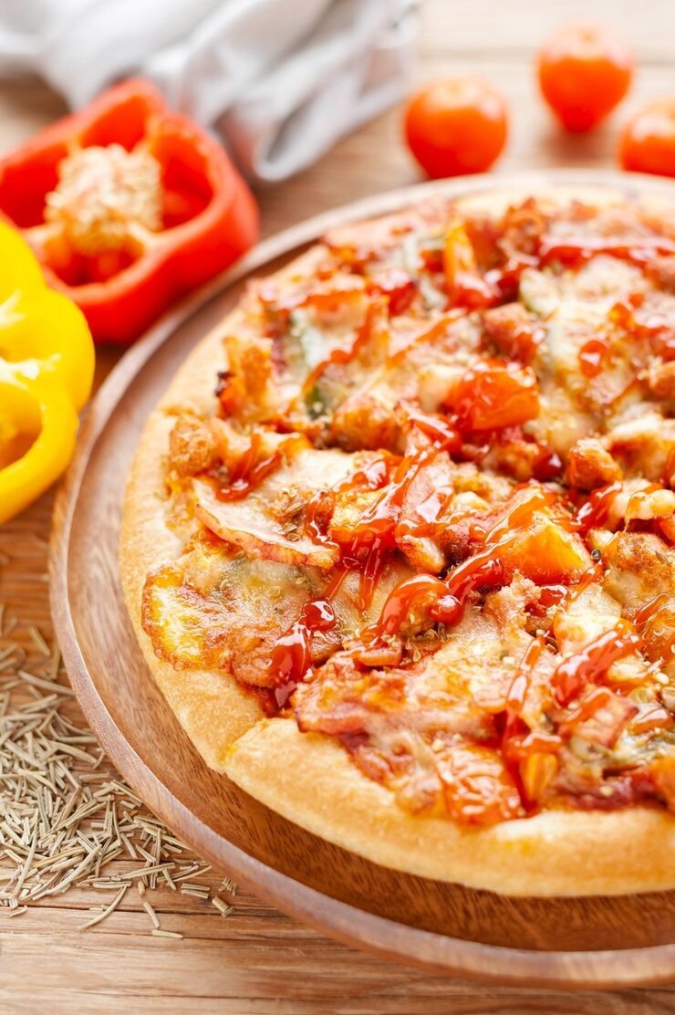 Close-up of a freshly baked pizza with melted cheese, pepperoni, chicken, and barbecue sauce on a wooden plate, with bell peppers and cherry tomatoes in the background.
