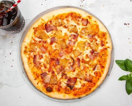 A pizza with cheese and bacon on a round metal tray, placed on a white surface, with a glass of soda with a straw nearby and green leaves in the corner.