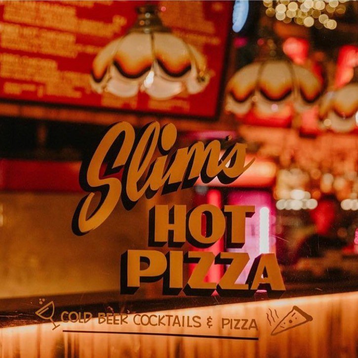 Sign advertising 'Slim's Hot Pizza' with a note about cold beer, cocktails, and pizza. The background features a cozy bar setting with dim lighting and vintage lamps.