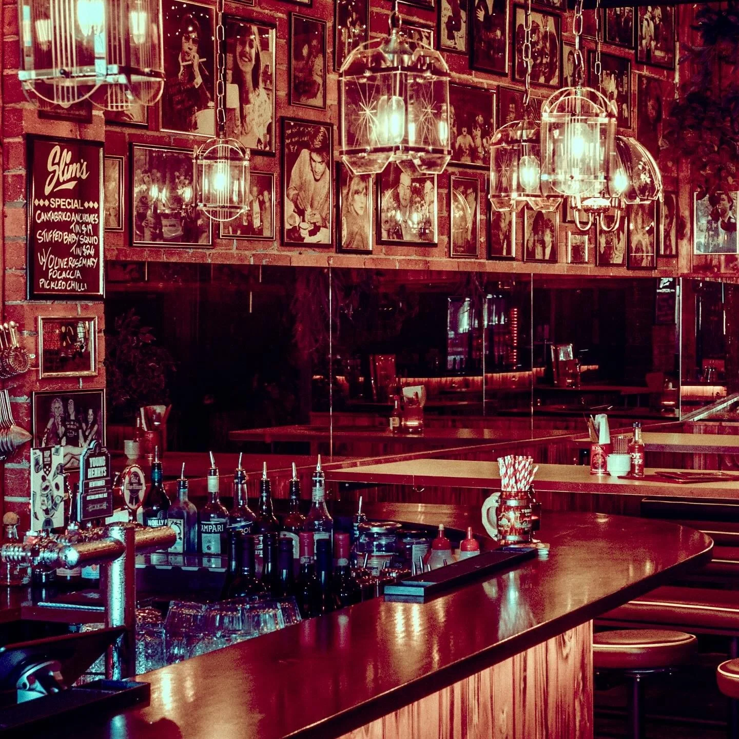 Interior of a vintage-style bar with a brick wall decorated with black-and-white photos. There are hanging light fixtures, a bar counter with various bottles and drink accessories, and mirrors reflecting the space.