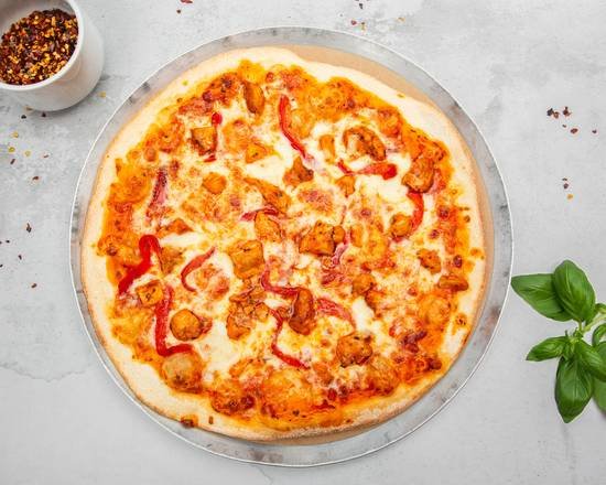 Pizza with cheese, chicken, and red peppers on a round tray, with a bowl of red pepper flakes and a basil plant nearby on a light grey surface.