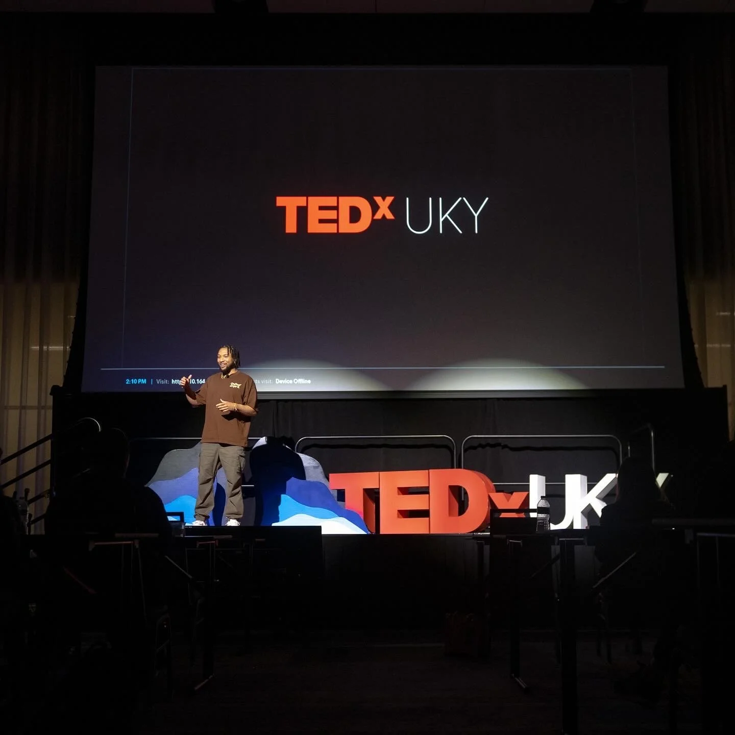 A speaker on stage at a TEDx UKY event, with a large screen behind displaying TEDx UKY logo, and large 3D TEDx letters on the stage.