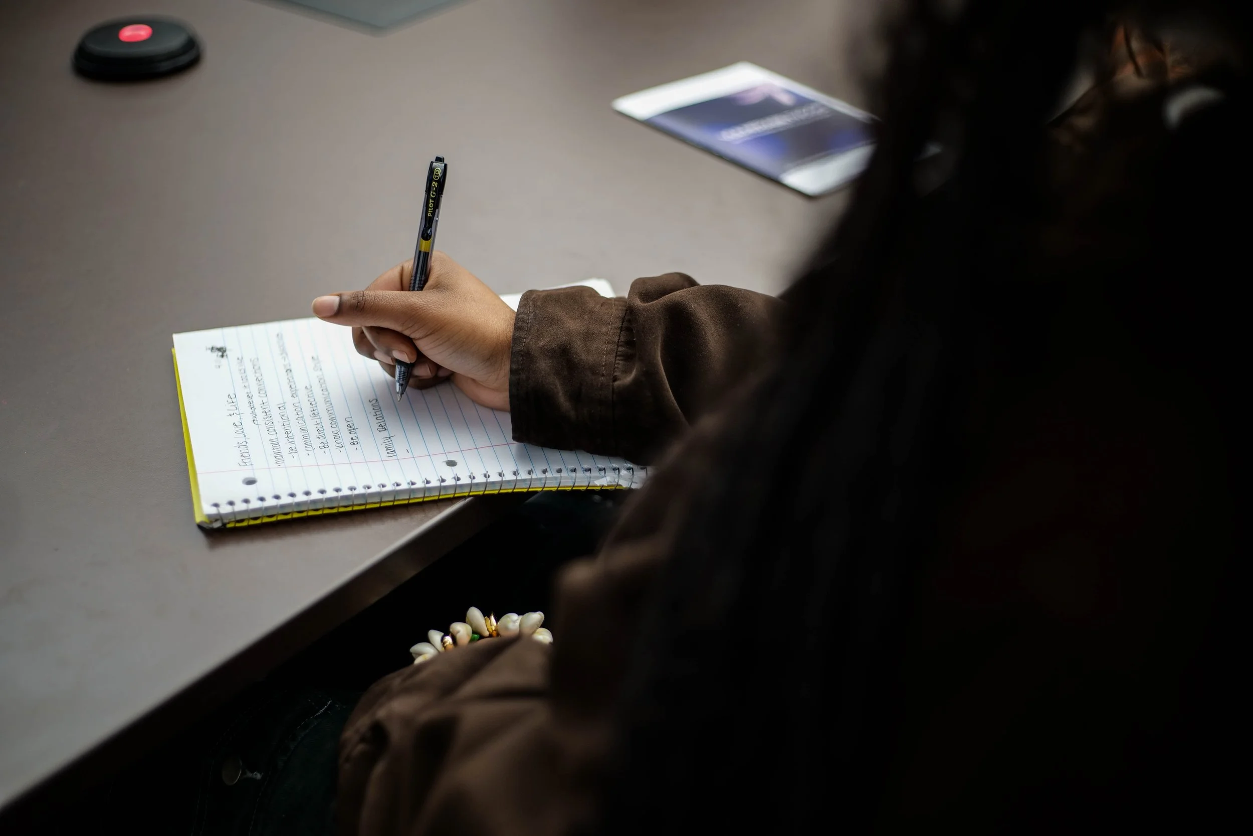A person writing notes in a spiral notebook with a black pen, sitting at a table with a blurred name badge and online meeting device nearby.