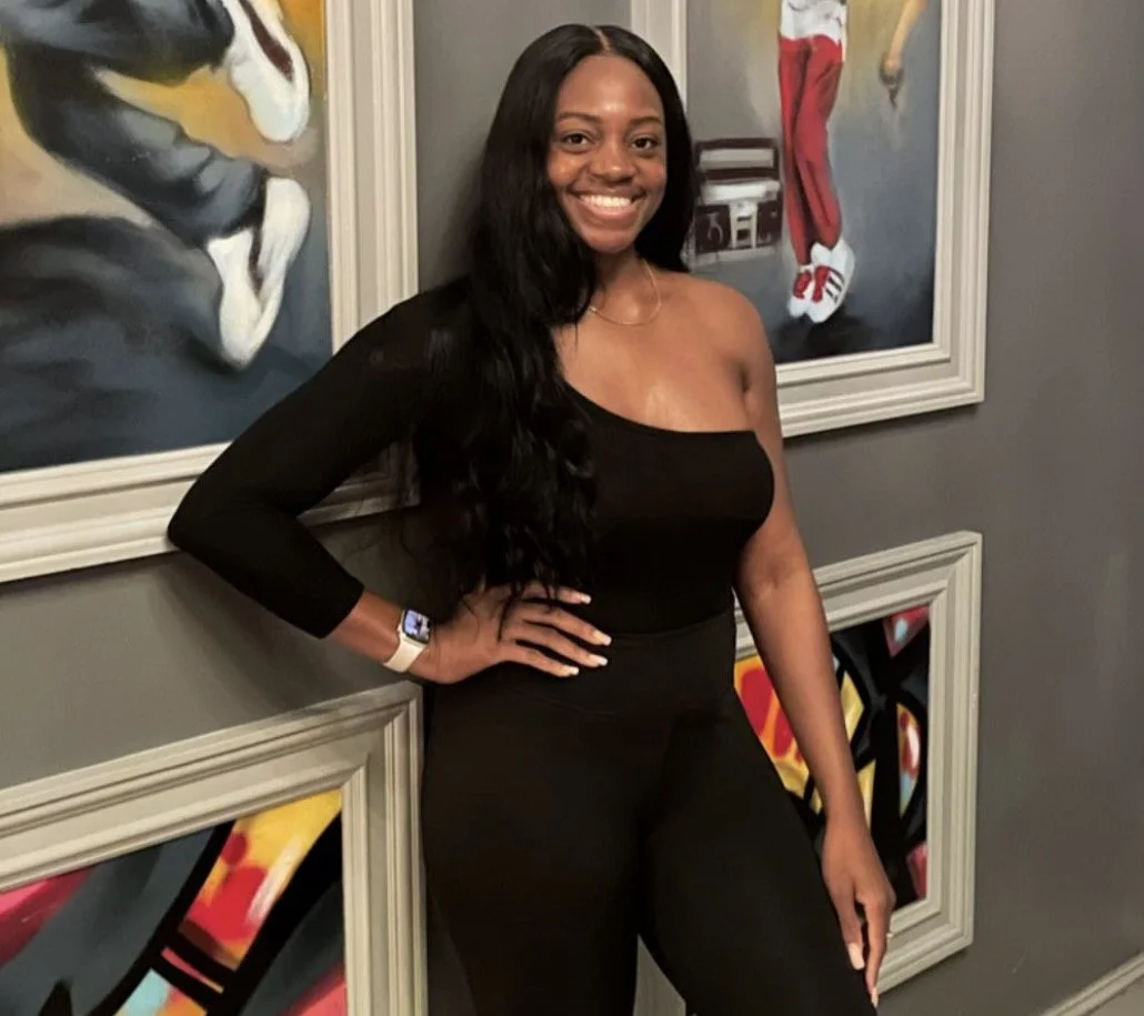 A smiling woman with long black hair wearing a black off-shoulder top and black pants standing in front of colorful abstract art on a gray wall.