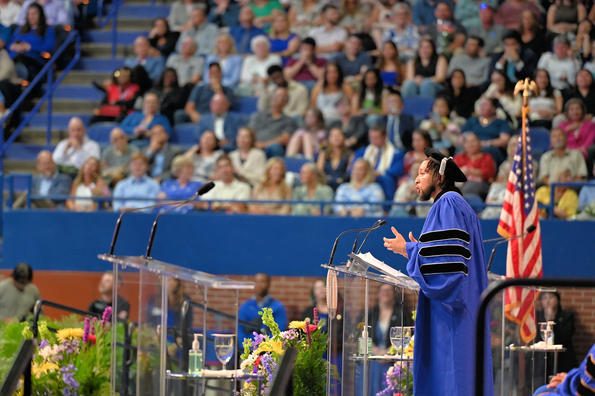 A man in a blue graduation gown and cap speaking at a podium with the American flag behind him during a graduation ceremony in an indoor stadium filled with seated audience.